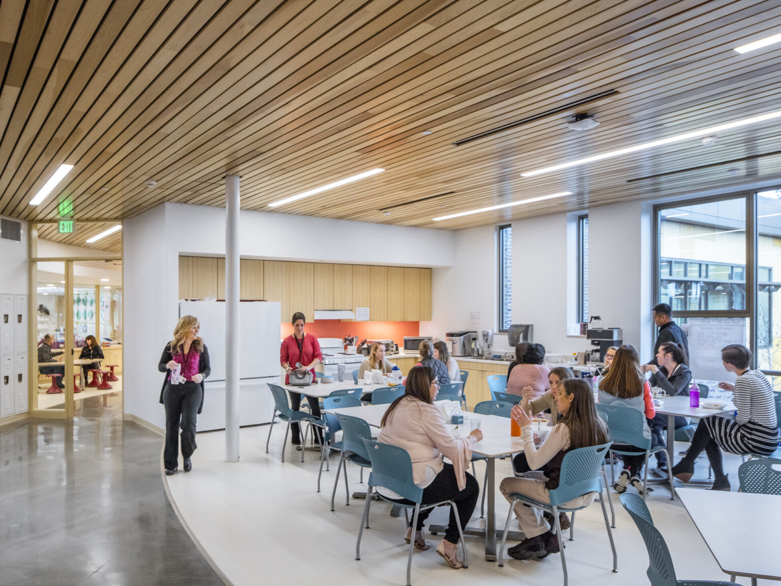 Workplace breakroom filled with tables and chairs with natural wood cabinets lining a back wall and floor to ceiling windows on the far wall