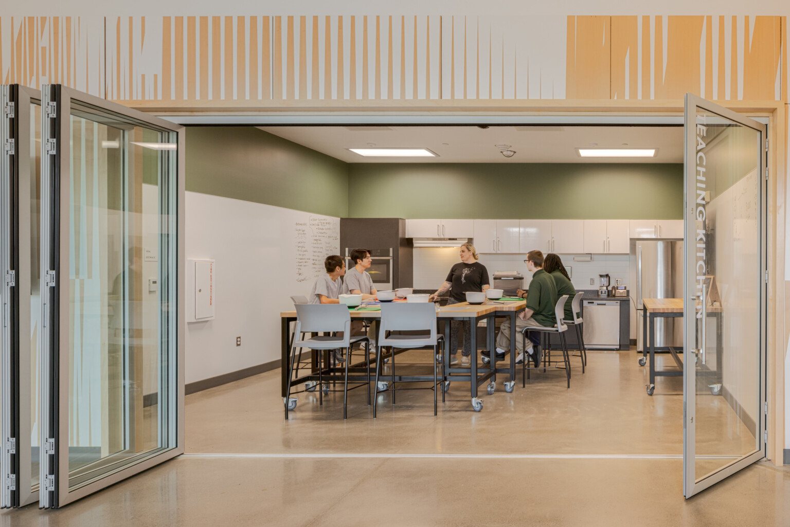 Teaching kitchen filled with students sitting at a high table in front of a wall lined with white cabinets and appliances