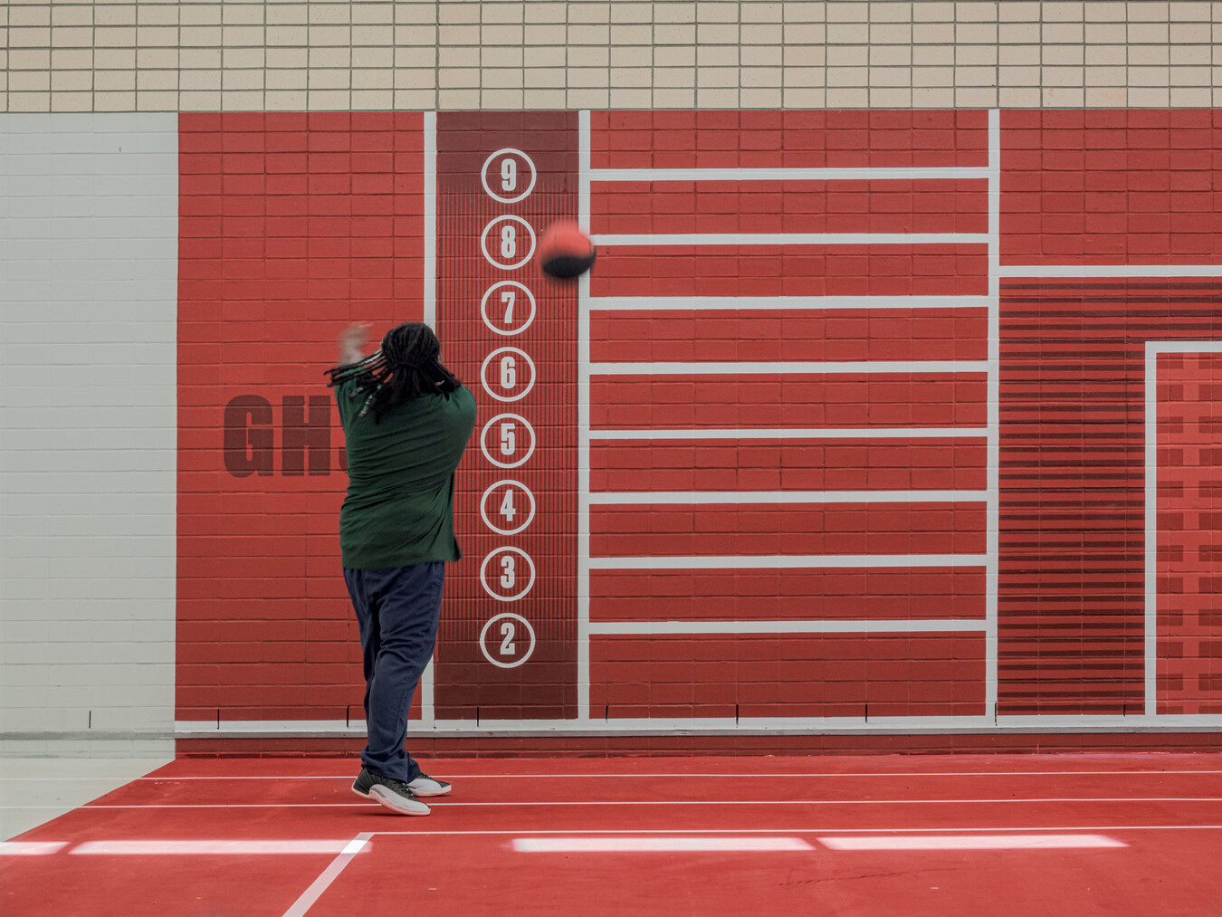 Kid in a green shirt throwing a call at a red wall with numbers 2 through 9 and lines