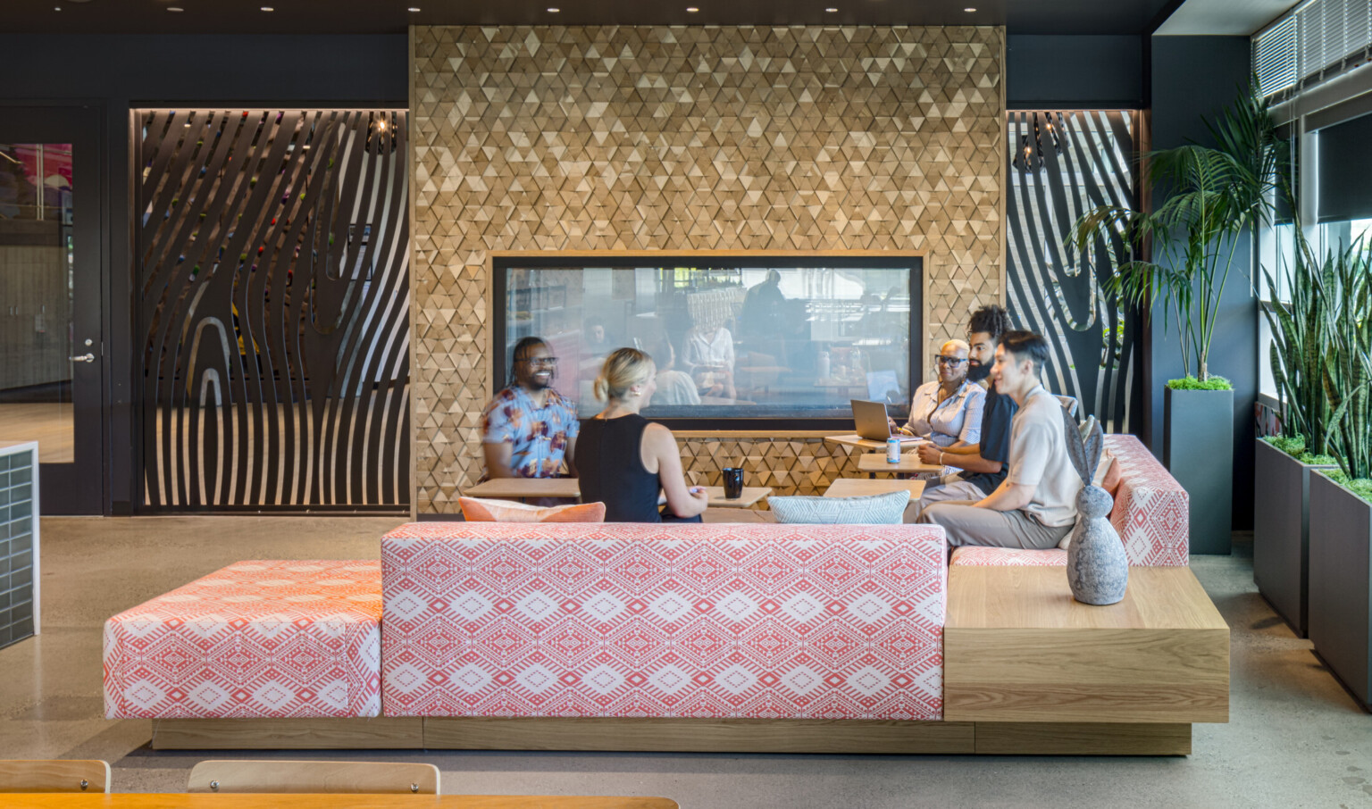 workplace lounge with a group of people gathered in front of a modern gold tiled fireplace. Modern furnishings with red and white print, light wood accent pieces, black walls