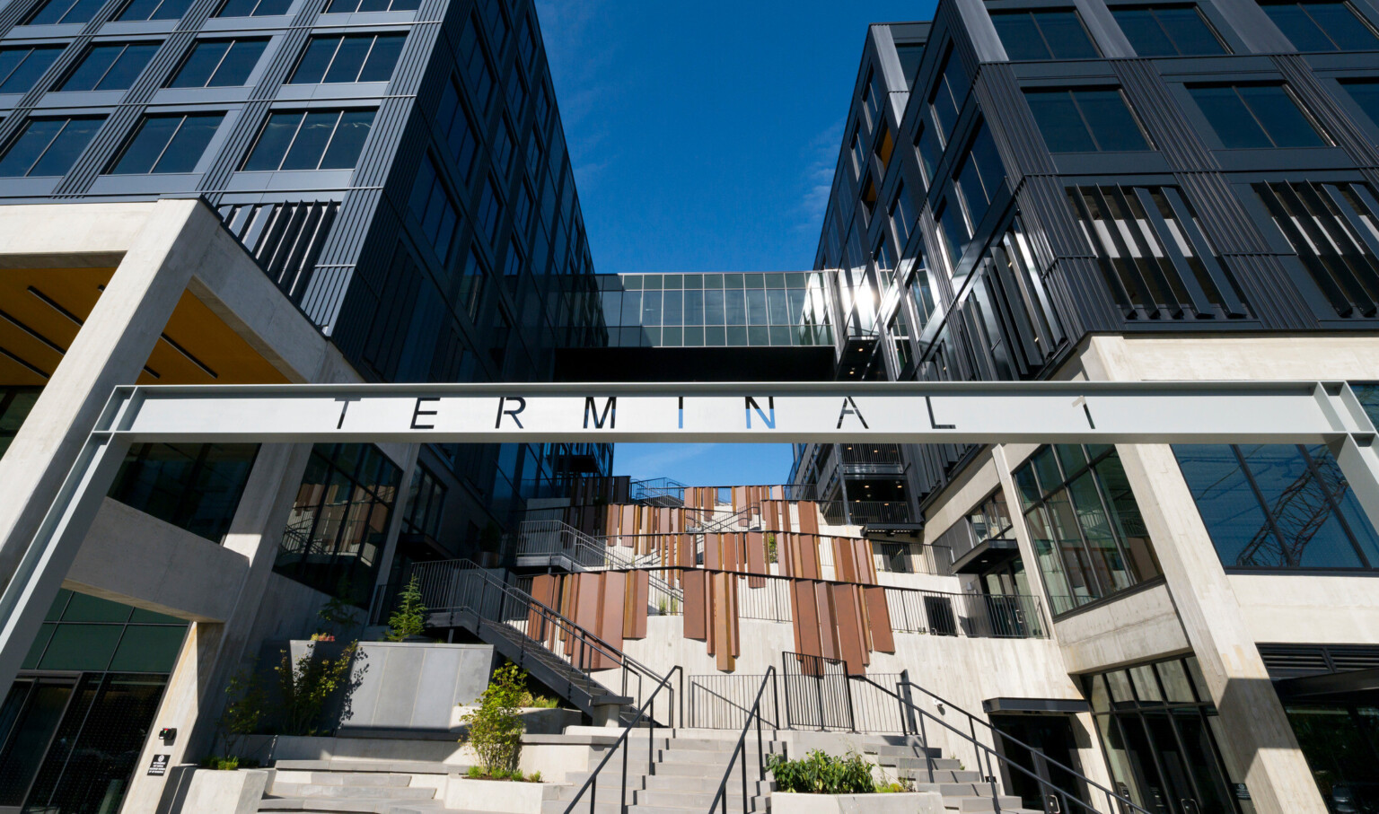 Two modern buildings flanking a central staircase. An enclosed glass walkway bridges their upper floors. A sign with large letters spelling "TERMINAL" hangs prominently across the middle of the image