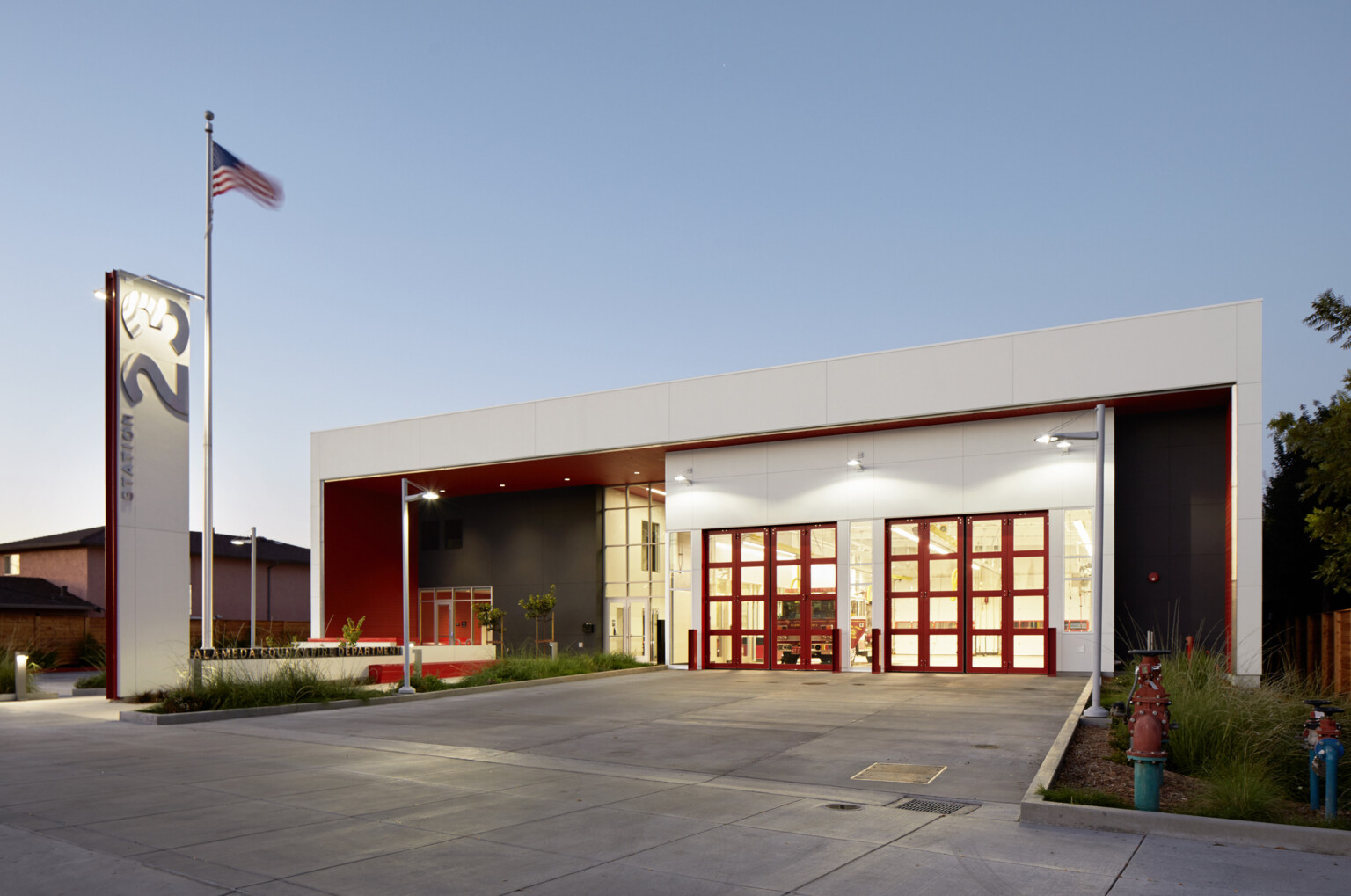 Front view of fire station and double-height entry portal. Two engine bays, a landscaped walkway. White and black façade with red accents.