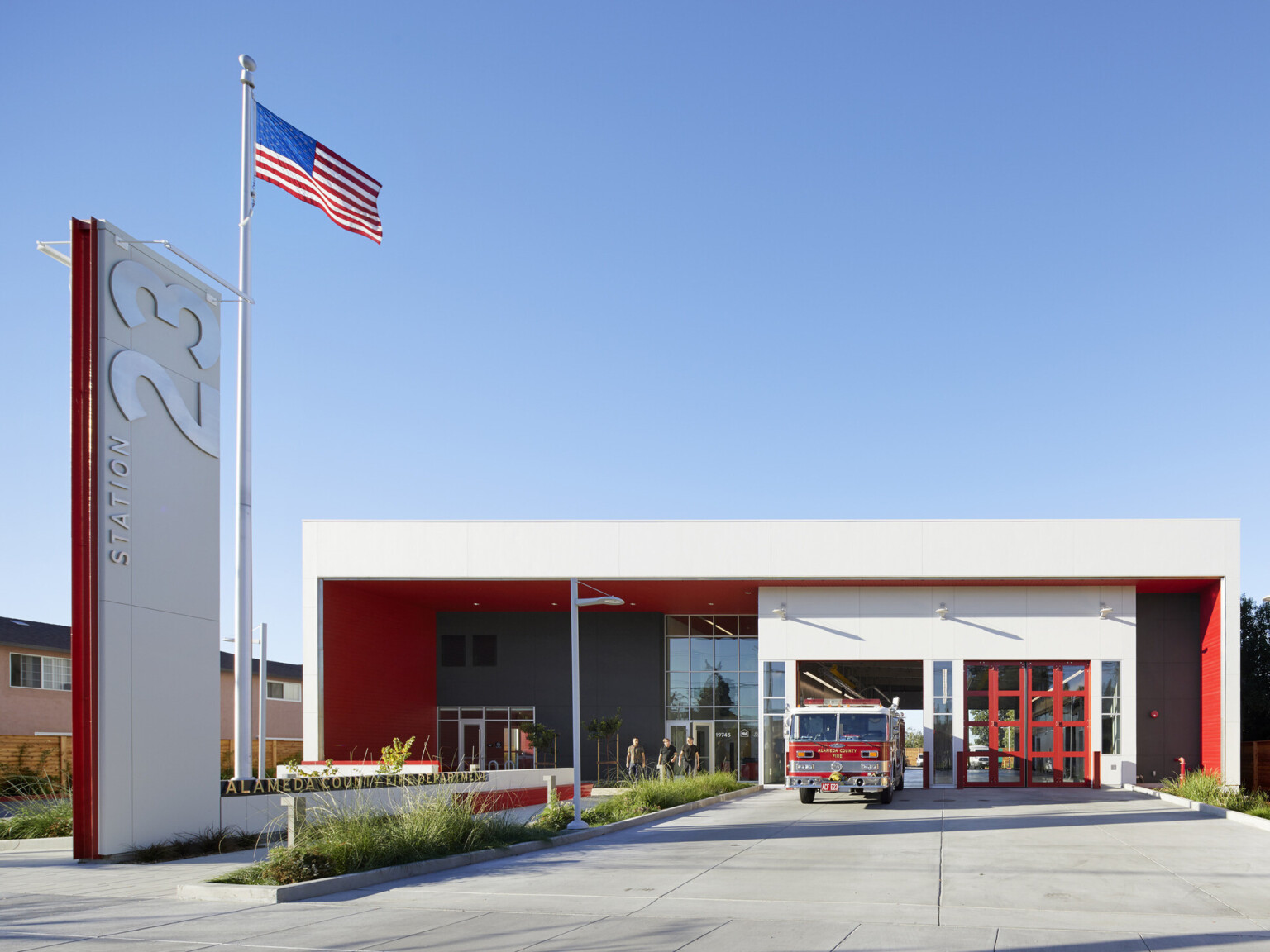 Front view of fire station with two engine bays, a landscaped walkway. White and black façade with red accents and a tall sign out front with station 23