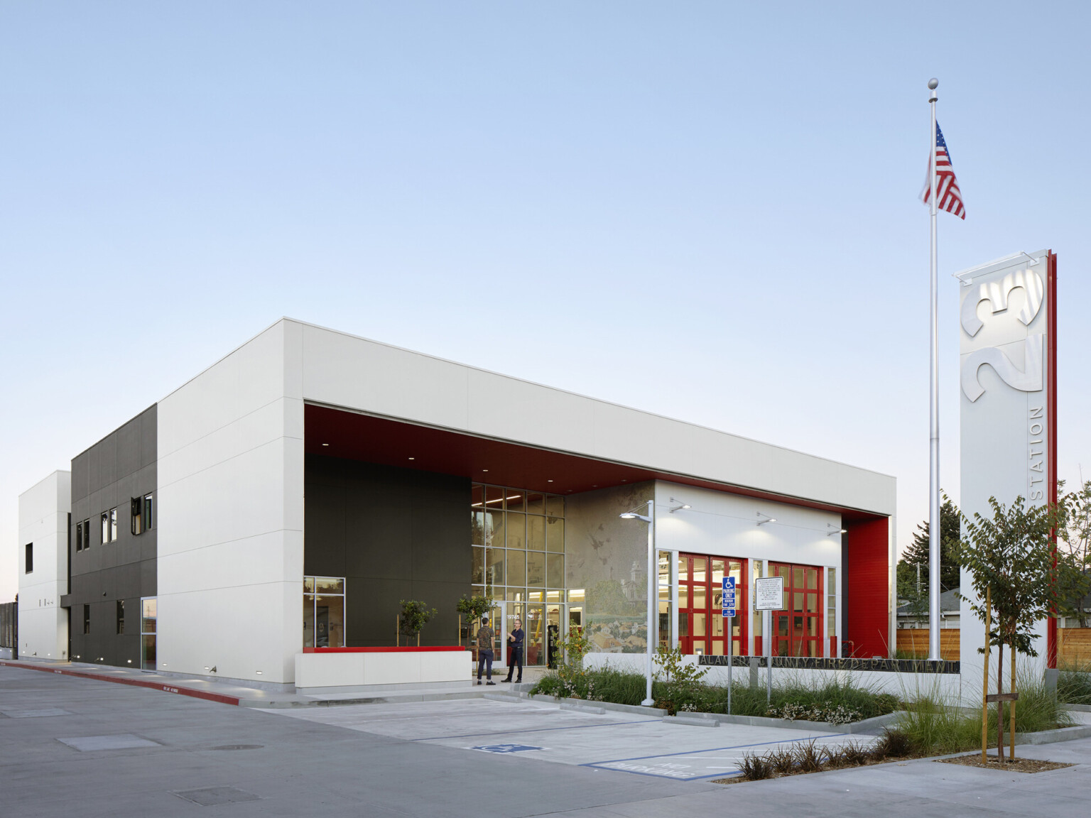 Side angle of fire station with two engine bays, a landscaped walkway. White and black façade with red accents and a tall sign out front with station 23