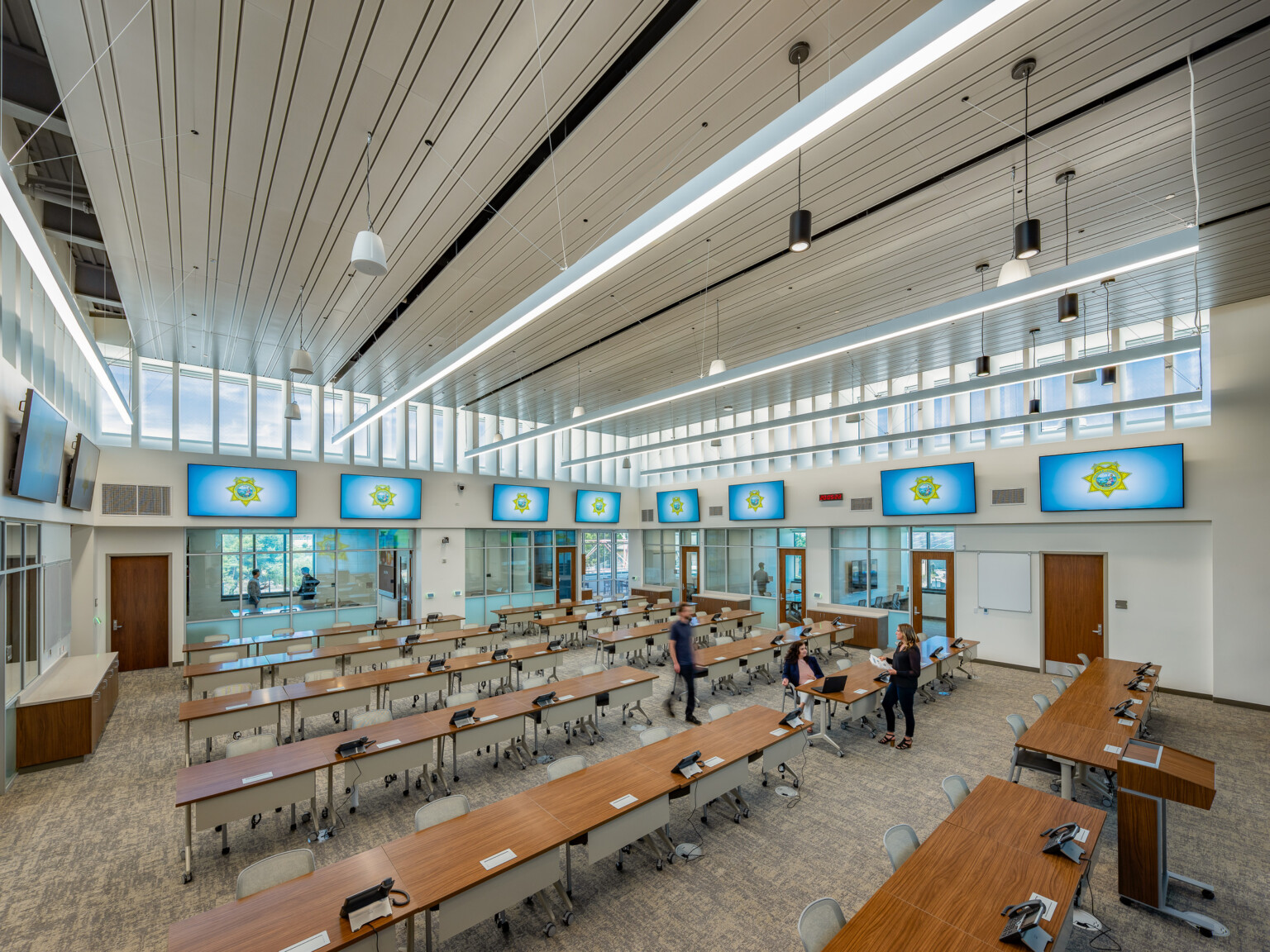 Conference room surrounded by windows filled with wooden top tables in rows with TVs in rows on top of the walls