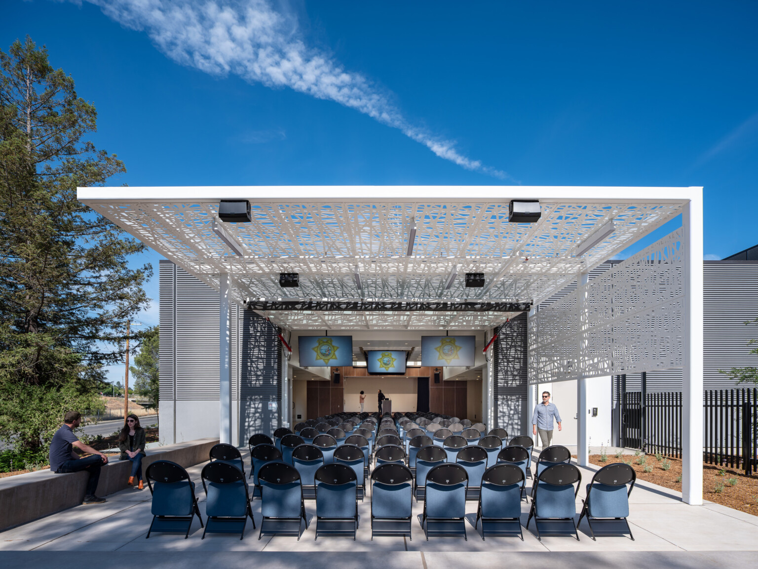 Exterior courtyard filled with chairs covered by a modern white awning looking at a podium with TV's suspended from the ceiling