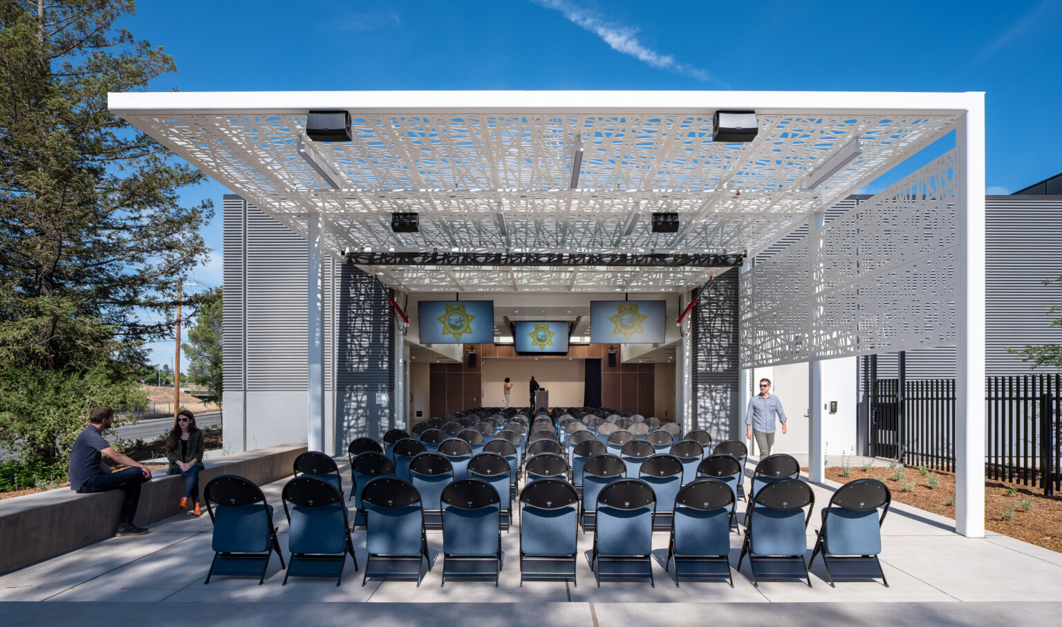 Exterior courtyard filled with chairs covered by a modern white awning looking at a podium with TV's suspended from the ceiling