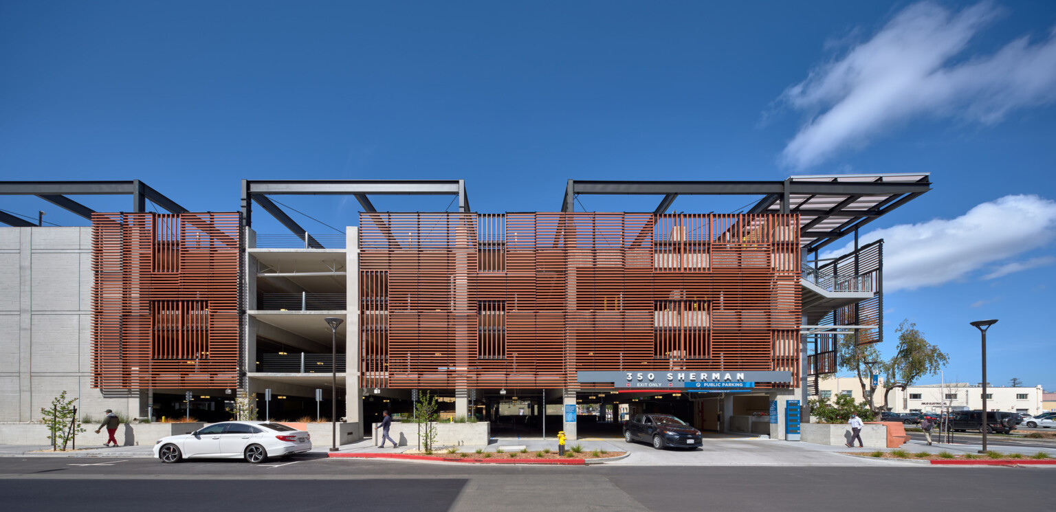 View of Palo Alto California Avenue Parking Garage entry with terracotta vertical screen system, precast panels, and exposed steel beams, car exiting and pedestrians walking along the sidewalk