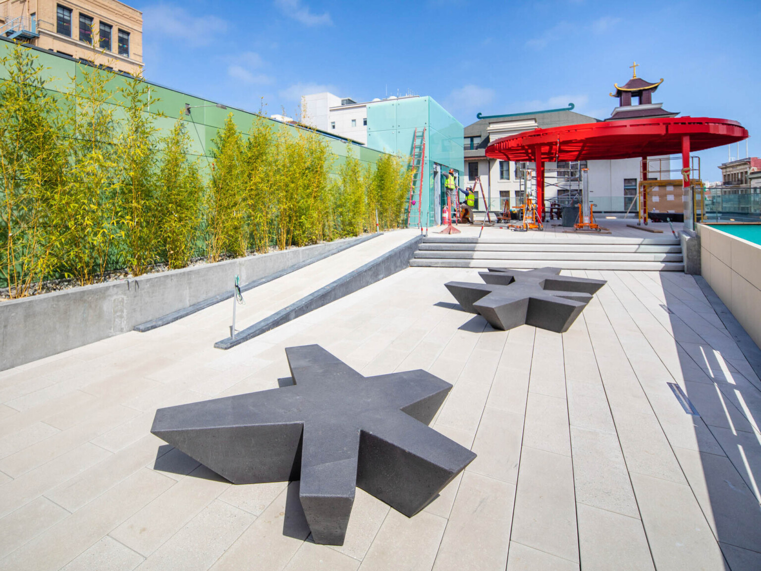 Organic shaped benches in walkway with ramp and steps to rooftop plaza with red canopy. Trees grow along green tile left wall