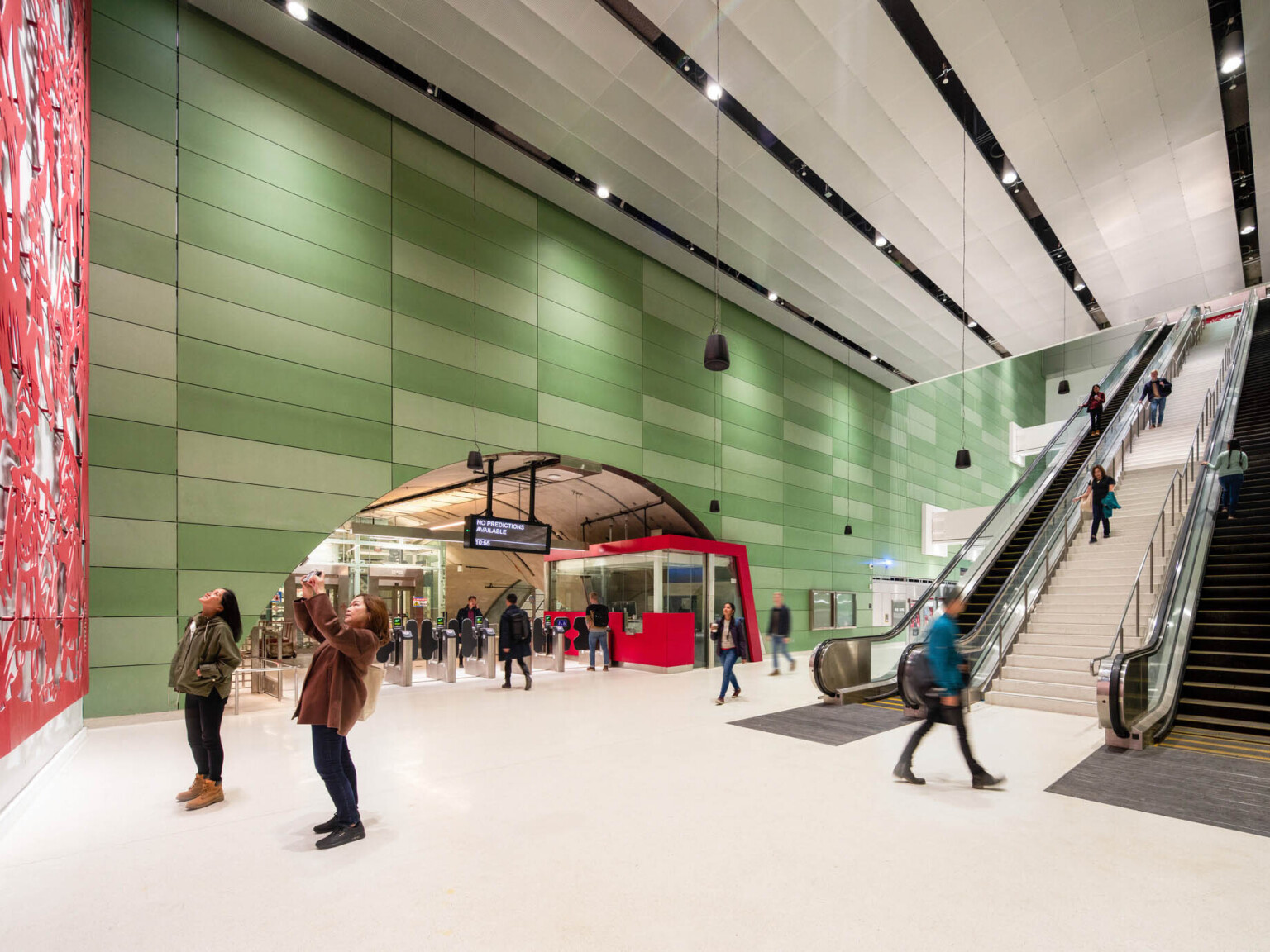 2 people looking at red art mural in a sub ground train station, escalators to back, rounded entry over turnstiles left