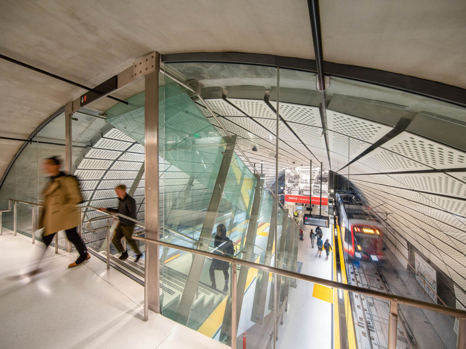 Rounded room with escalator to station below between 2 train tracks. White panel ceiling. Glass wall dividing tunnel