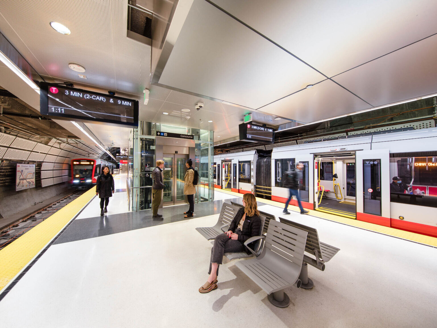 Rounded side walls of tunnel covered in perforated white panels. Hanging light beams along tracks beside screens, elevator at platform center