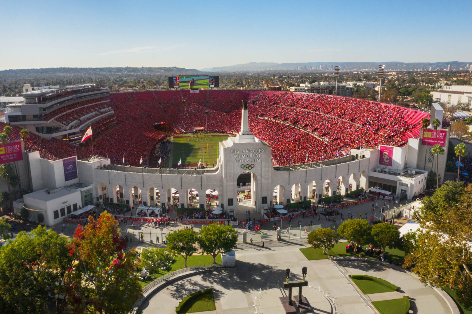 Front view of full stadium labeled Los Angeles Memorial Coliseum with Olympic Rings logo on central tower framed by archways