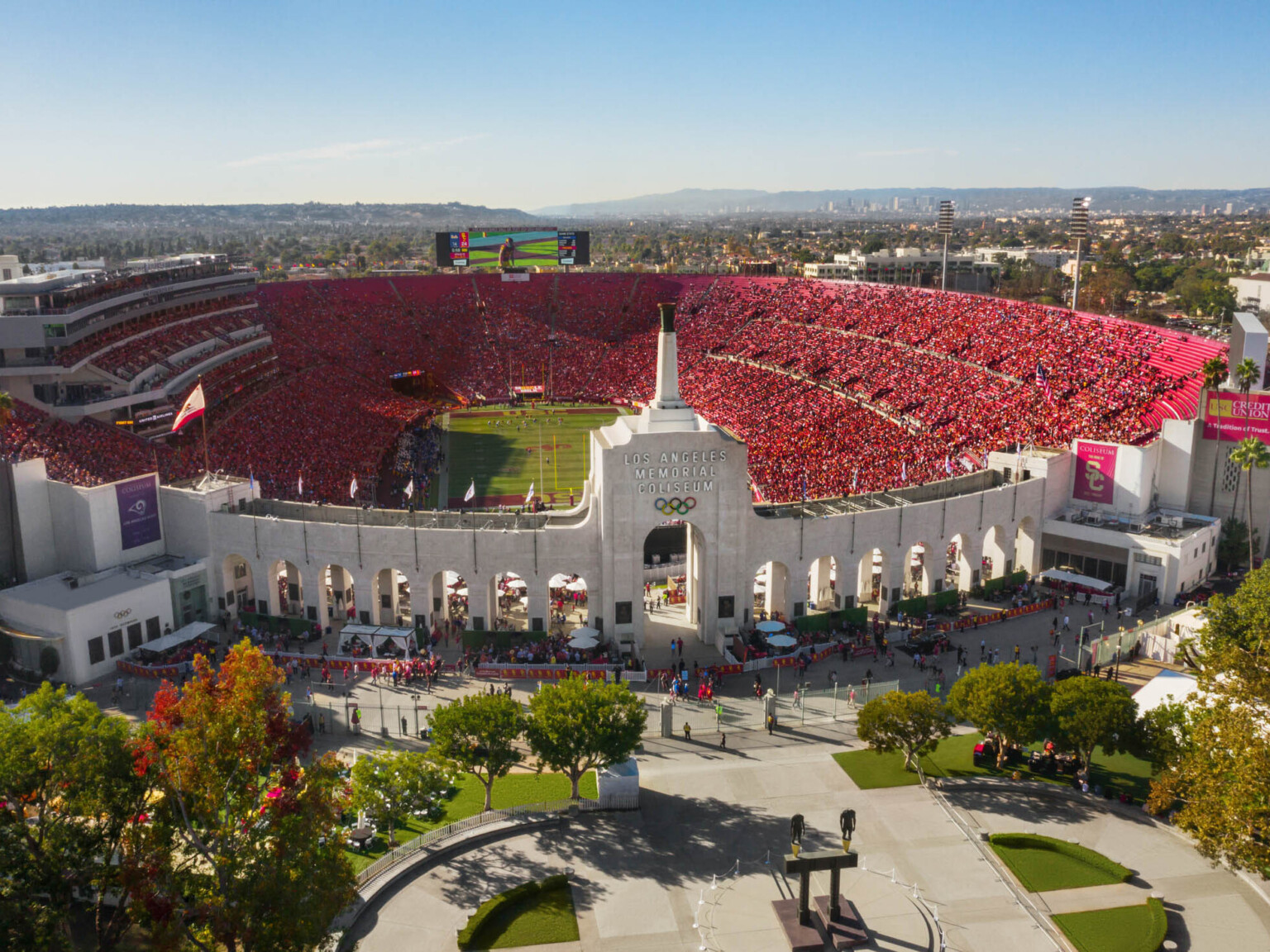 Front view of full stadium labeled Los Angeles Memorial Coliseum with Olympic Rings logo on central tower framed by archways