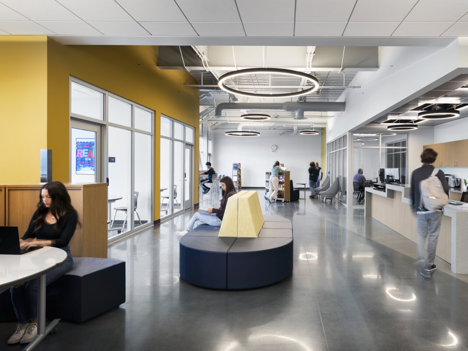 High school common area with modern circular lighting suspended from the ceiling filled with rounded seating and countertop to the back wall