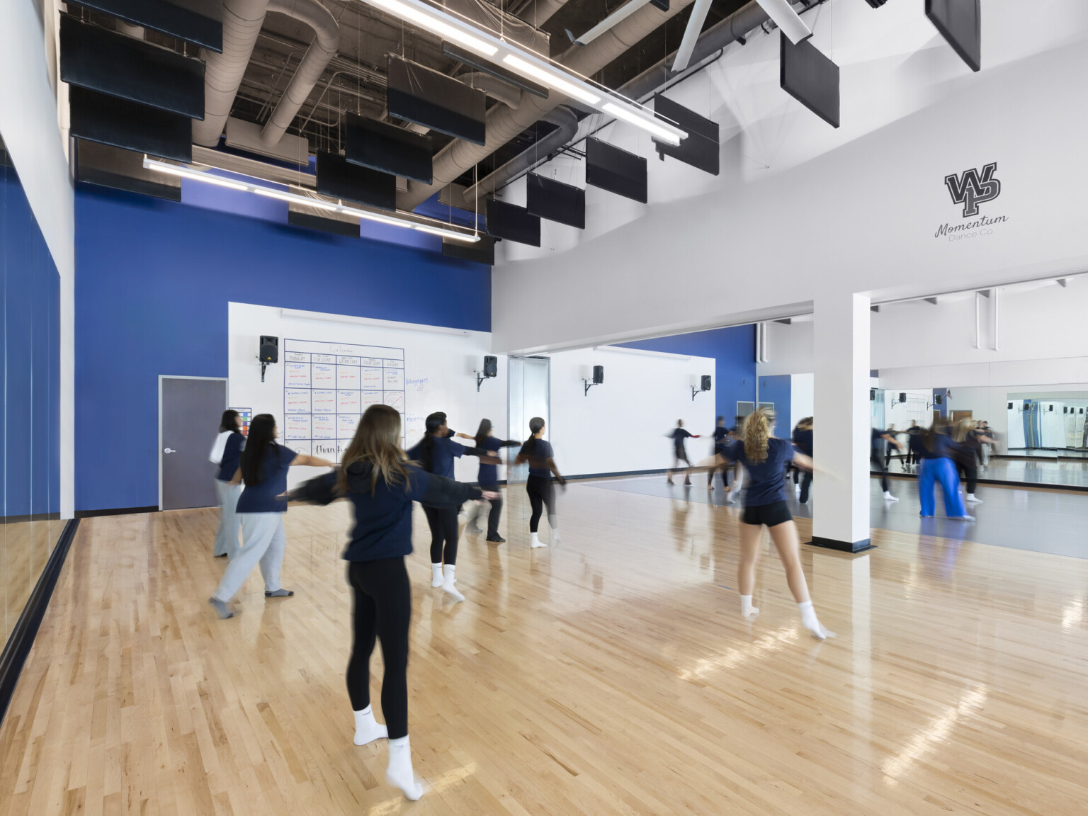 High school dance room filled with students. Blue accent wall adjacent to a wall full of mirrors