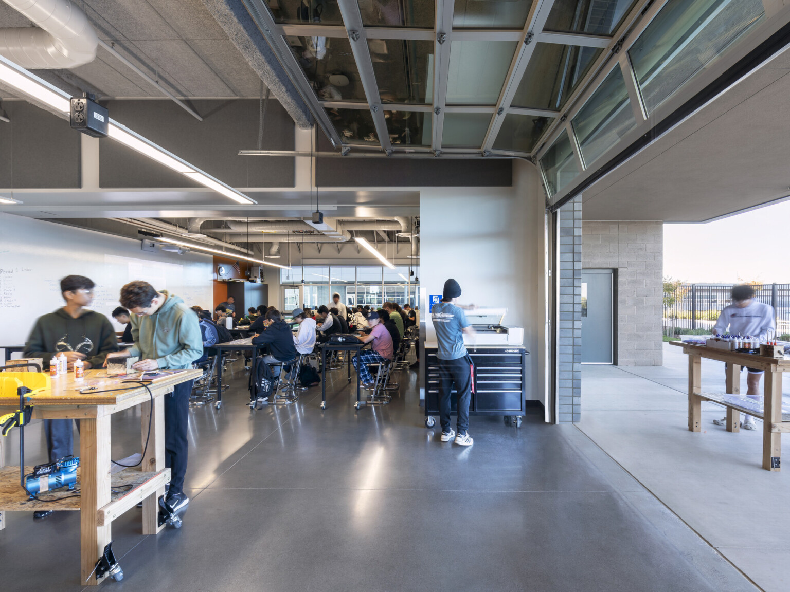 Classroom filled with students working at tables. Natural wood table in front of open roll up doors with more students outside