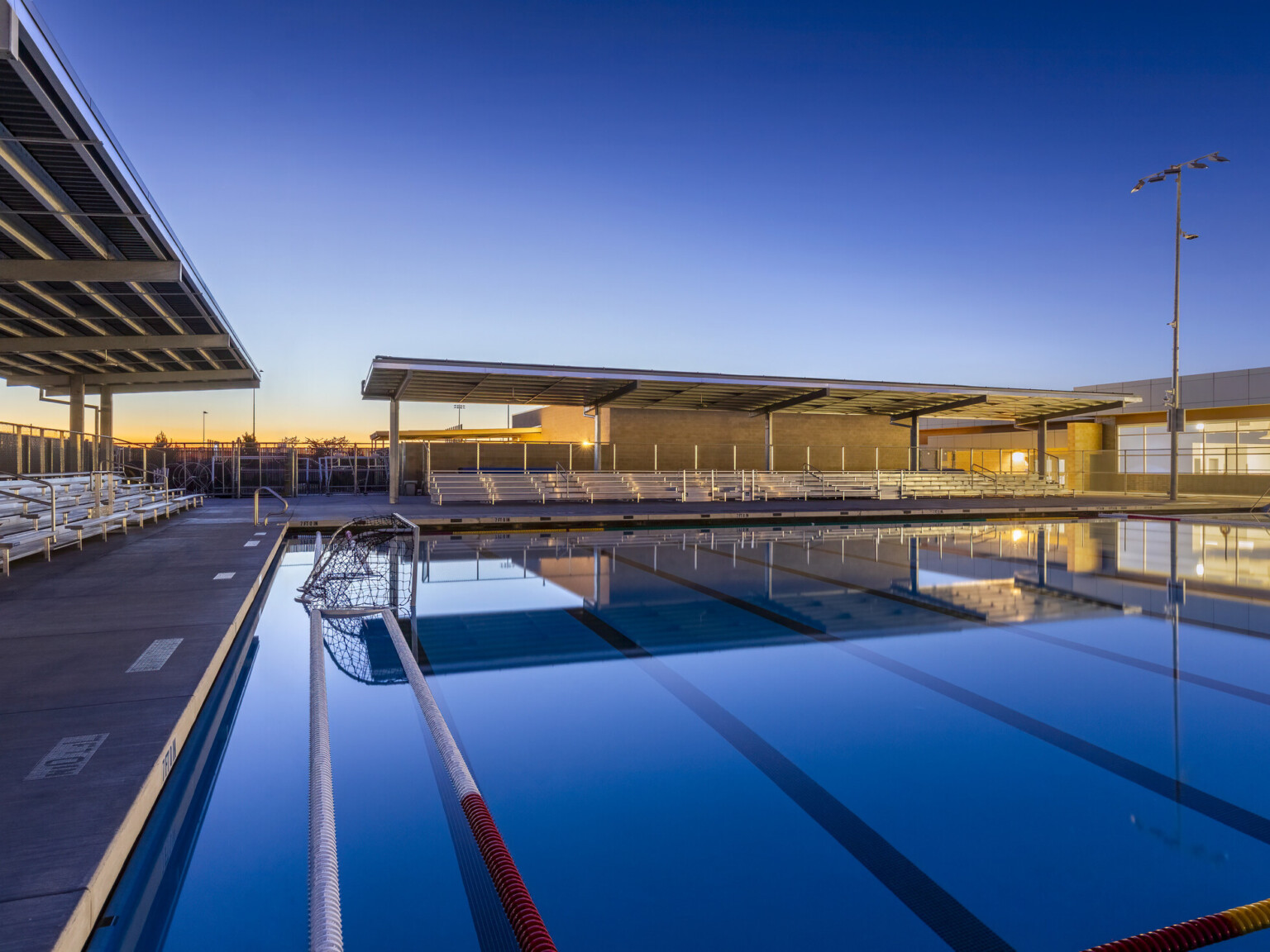 Outdoor swimming pool lined with bleachers at dusk in front of an illuminated building