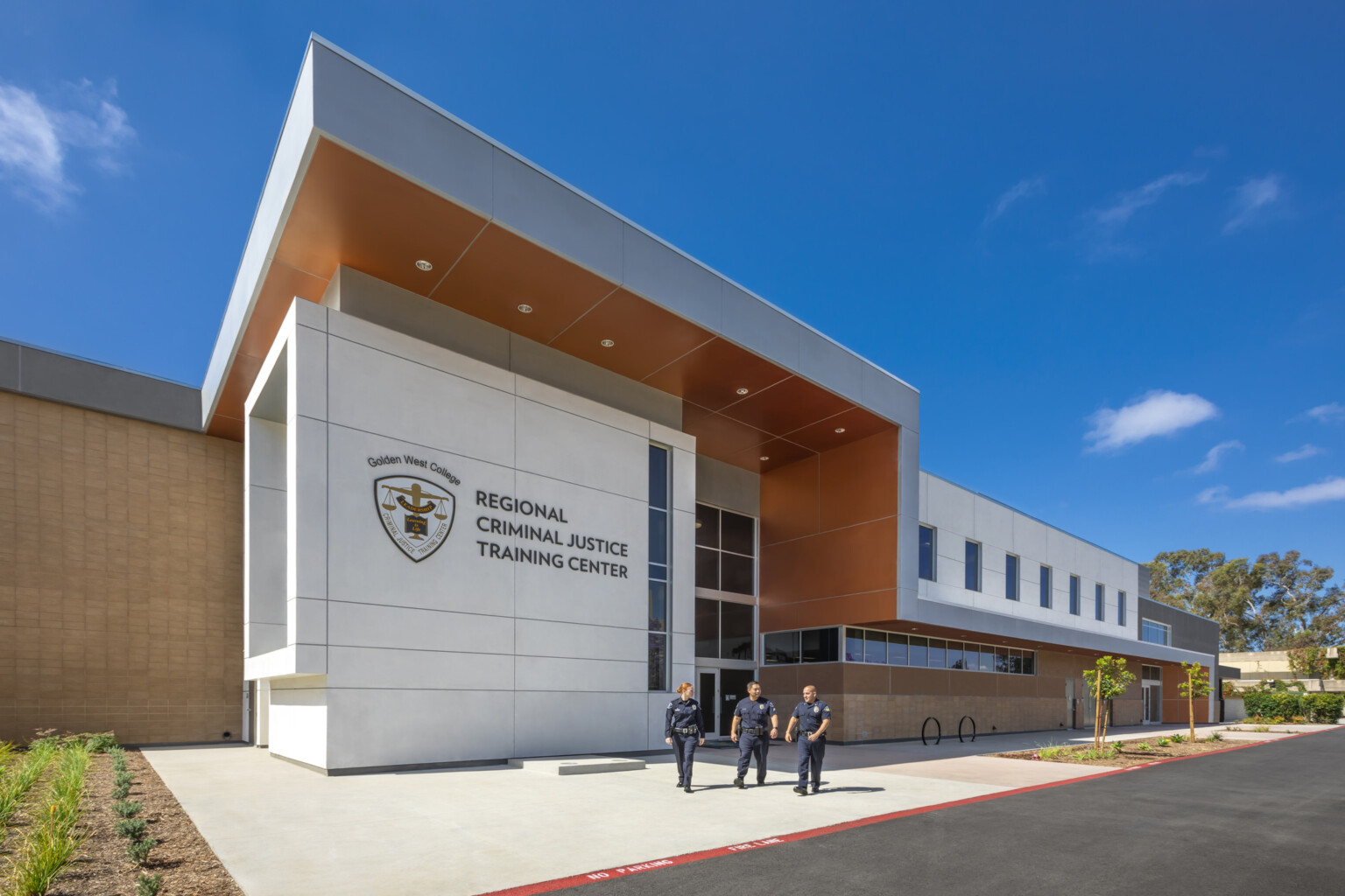 Corner exterior view of Golden West College’s Criminal Justice Training Center, a white cubic entrance with logo facing the street, shaded by a grey overhang with orange base