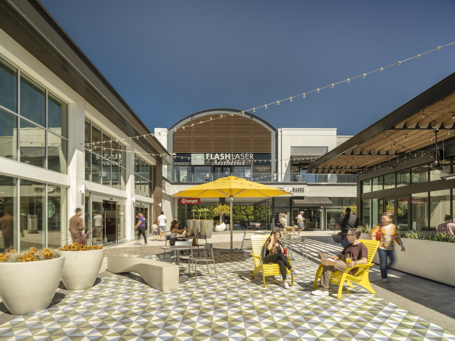 Courtyard in an outdoor shopping area surrounded by modern buildings with large glass windows, yellow chairs, a yellow umbrella, and geometric-patterned tiles