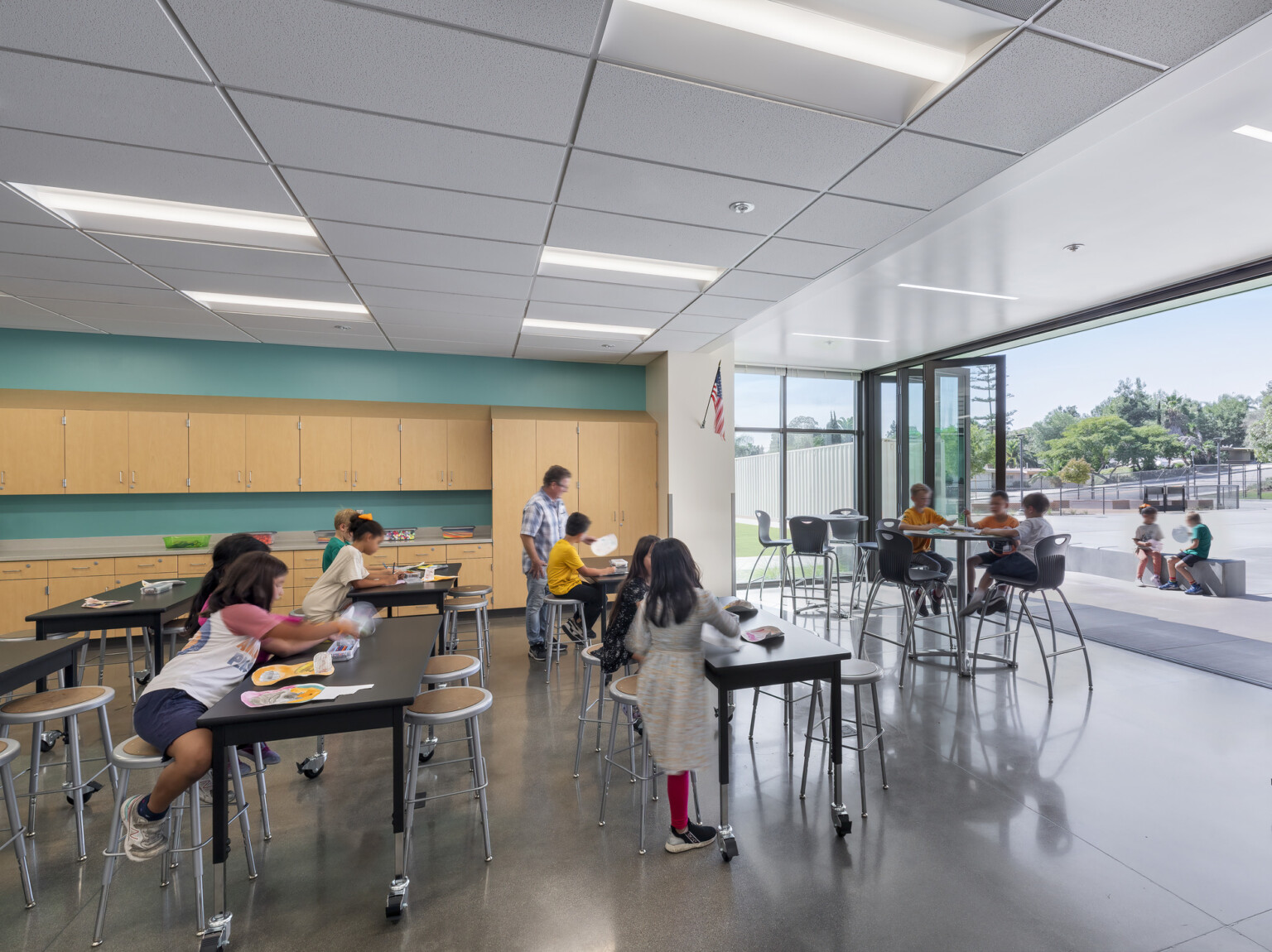 classroom with wood cabinets and mixed height seating spaces, floor to ceiling window wall opens to playground