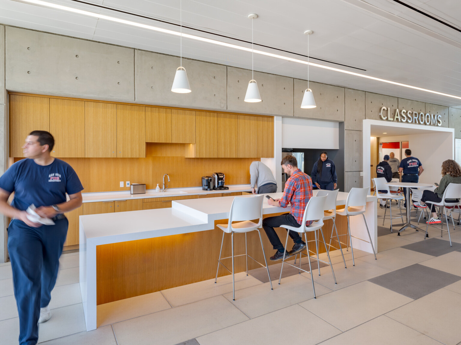 Break room with natural wood cabinets on a back wall behind a high top counter seating area. Multiple tables and seating areas to the right side in front of a hallway with word Classrooms on the wall