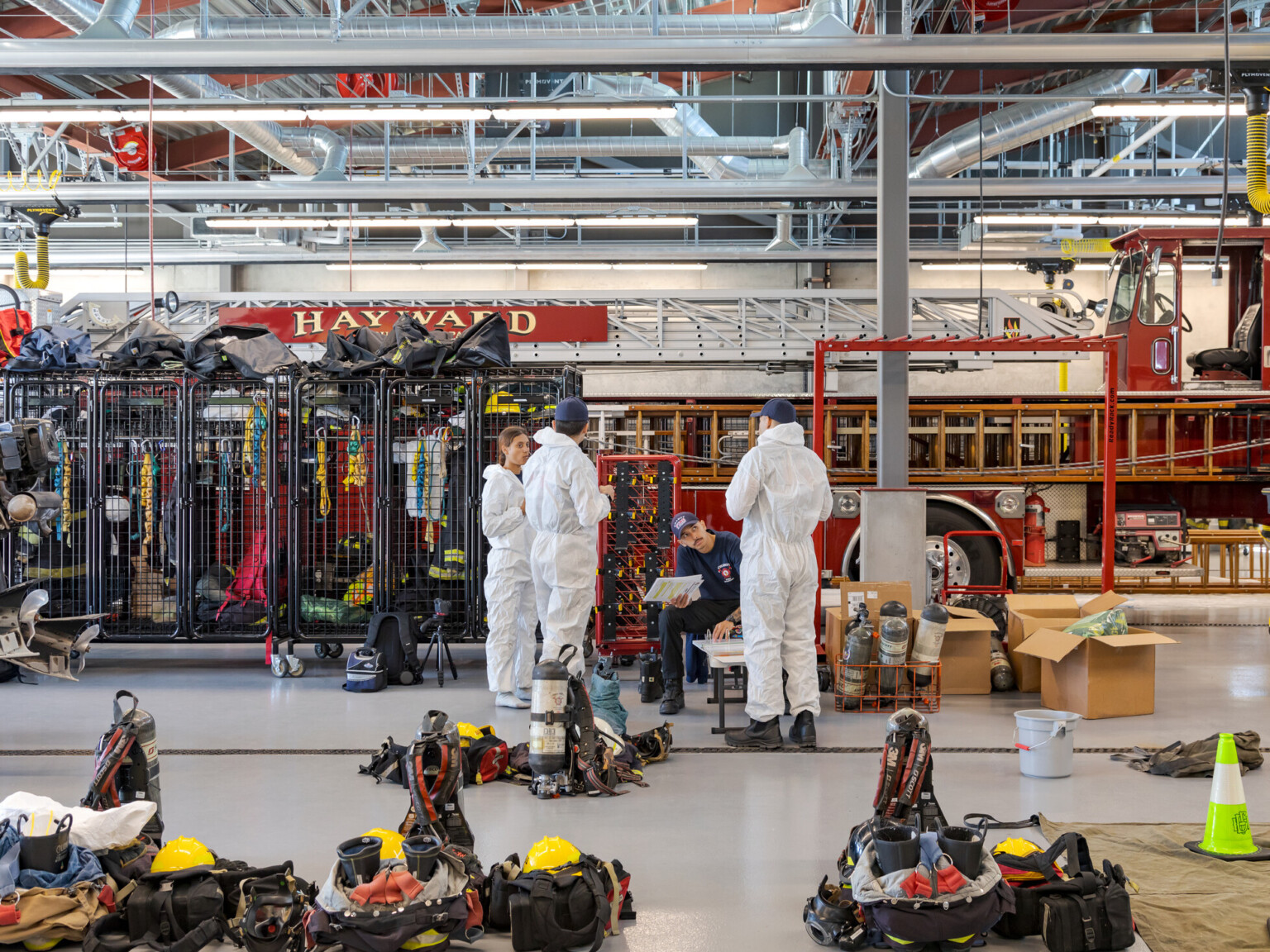 Group of fire department trainees in white suits standing in front of fire training equipment surrounded by safety gear