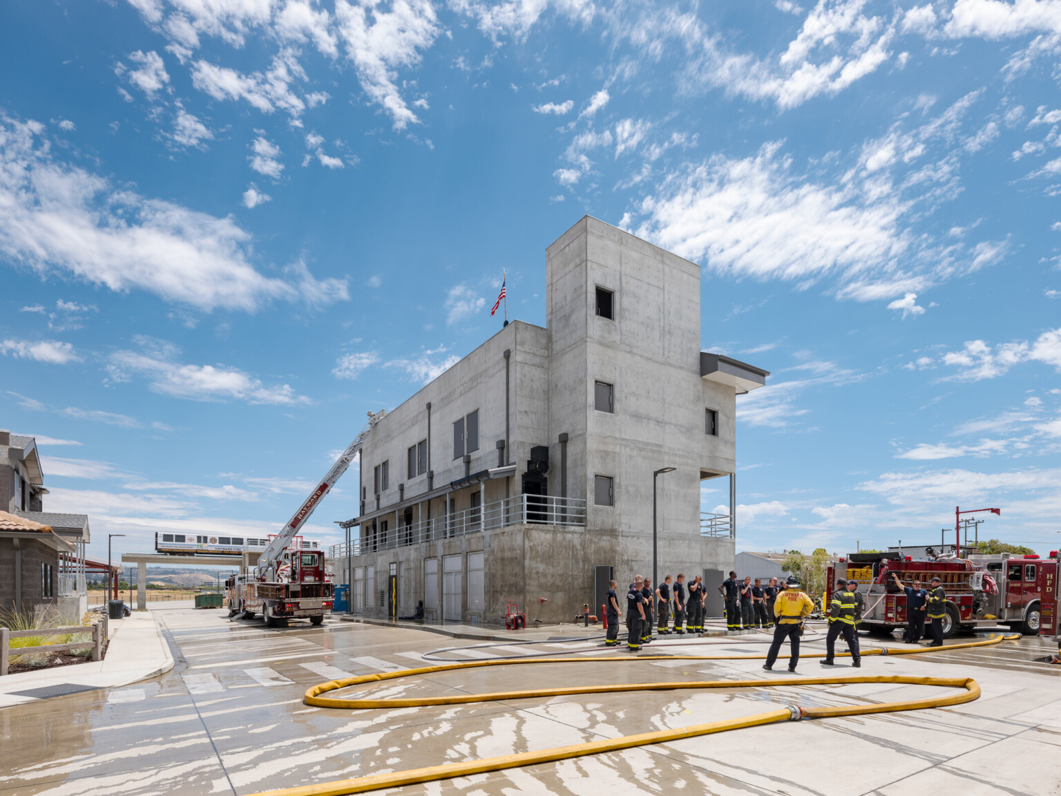 Firefighters standing in a semicircle standing outside of a concrete building with yellow hoses extended on the ground with multiple fire trucks in the image