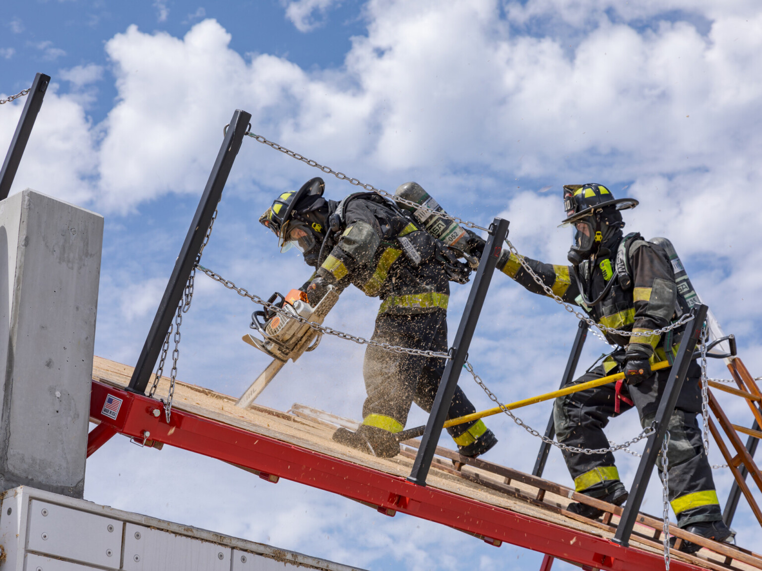 Two firefighters dressed in safety gear using a chainsaw to cut through a wooden floor