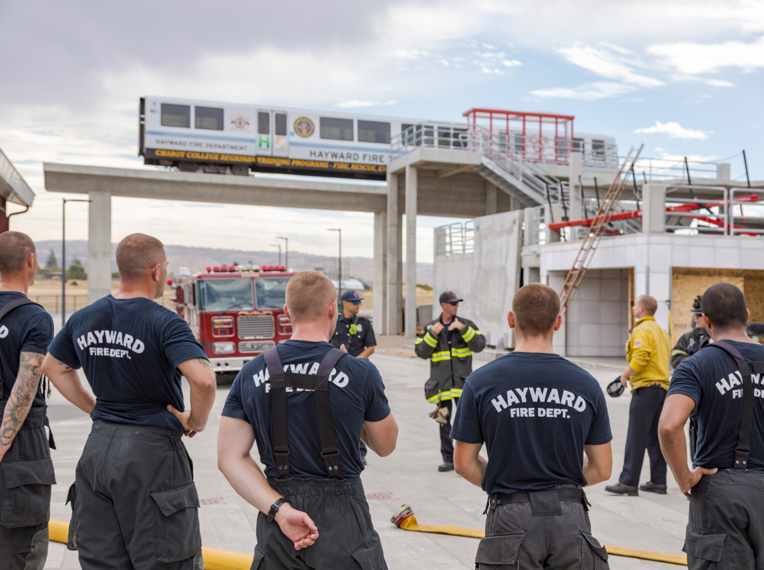 Firefighters in blue shirts with Hayward Fire Dept. on backs standing in front of a fire truck and building structure listening to instructors in safety gear