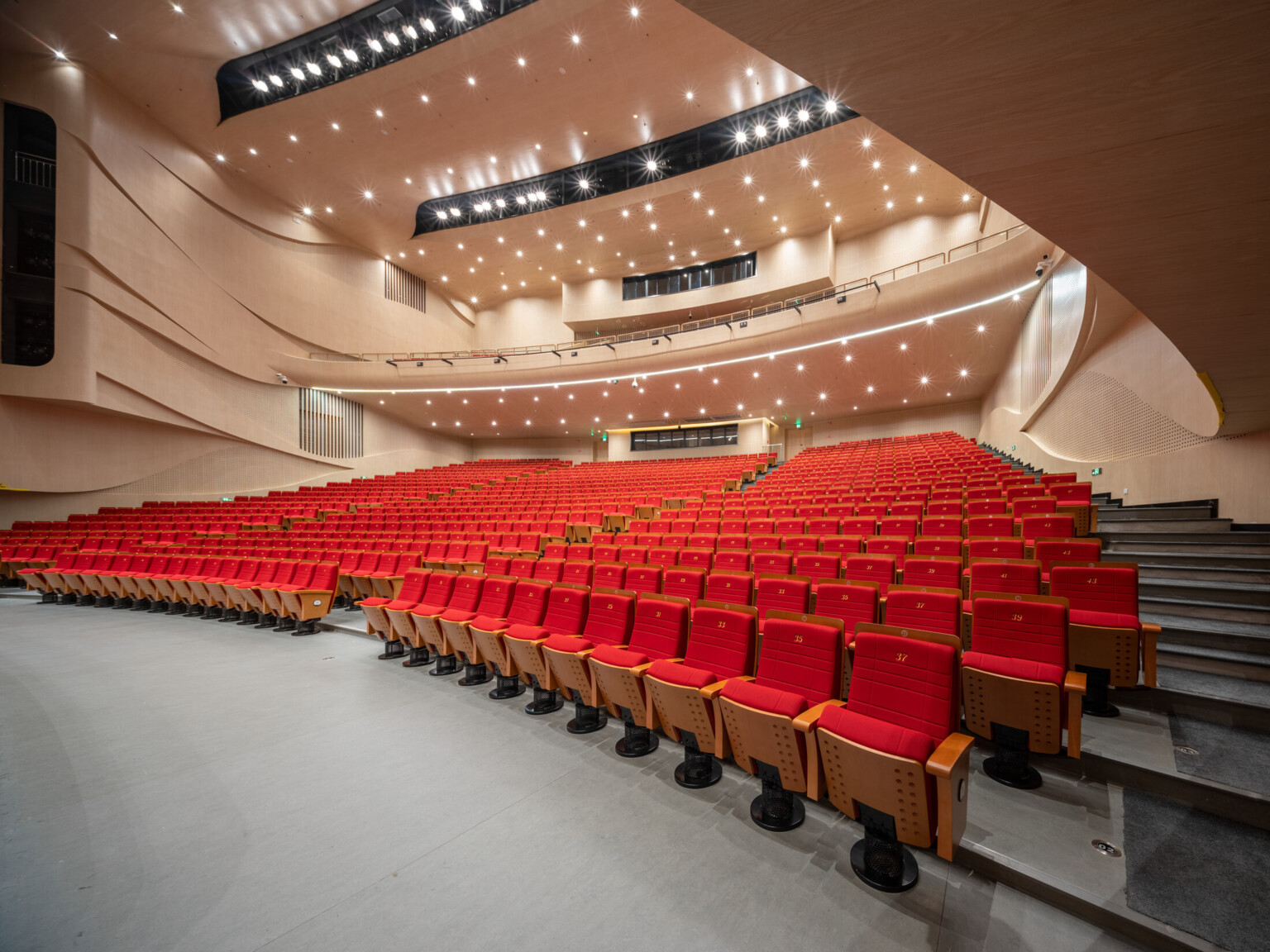 Multilevel theatre filled with red chairs with wooden arms and bottoms. Grey floors and beige walls