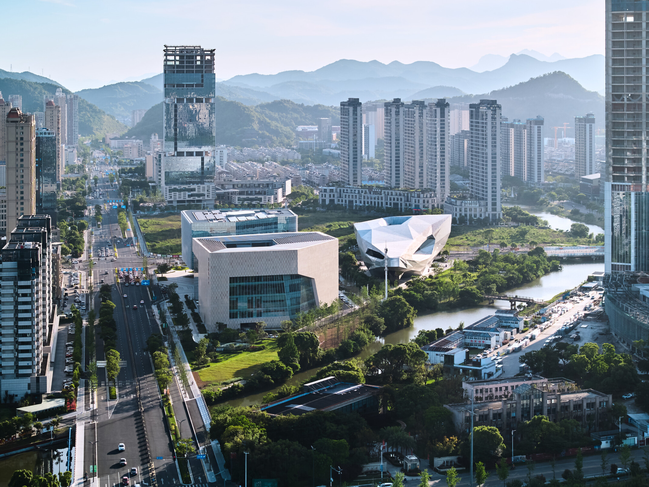 Aerial of the Wenling Cultural Center showing multiple multistory buildings along a river with a large roadway to the left and high rise buildings in the backdrop