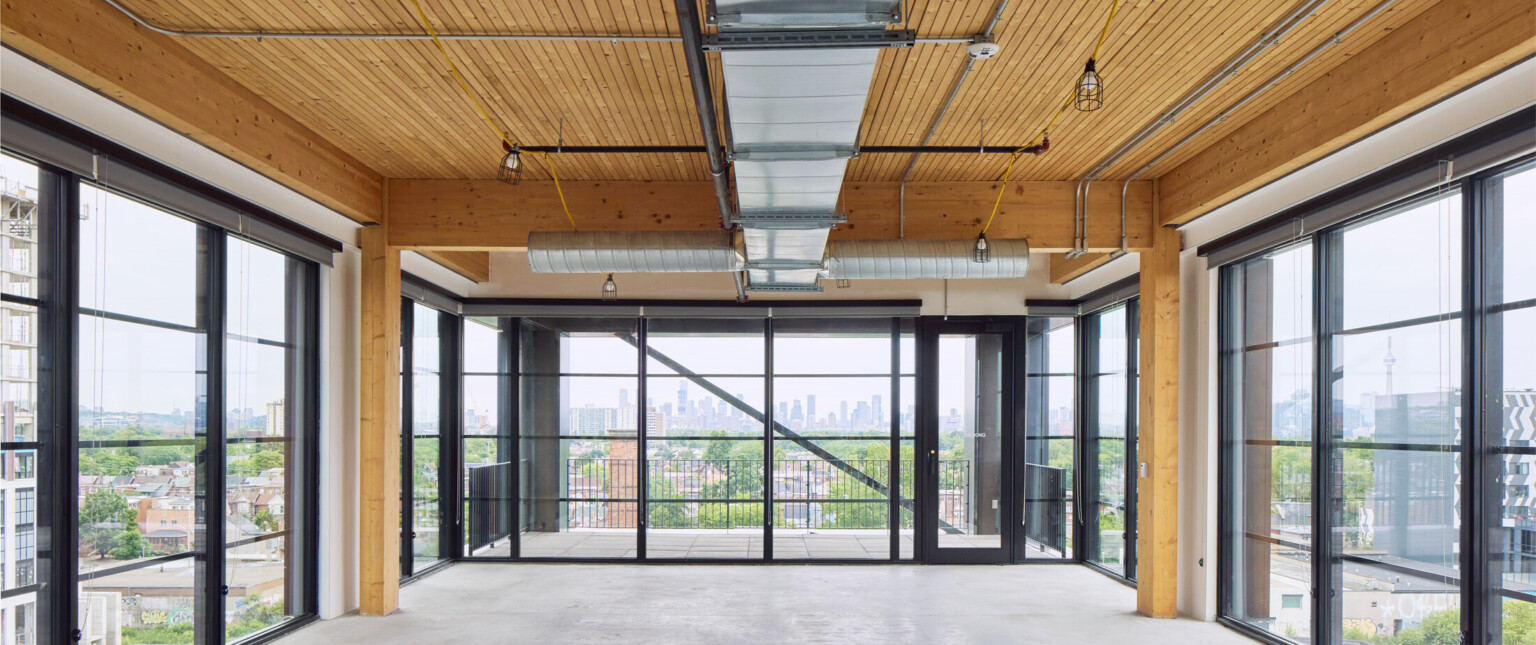 Large room with exposed wooden beams, floor to ceiling black framed windows overlooking a skyline filled with large buildings