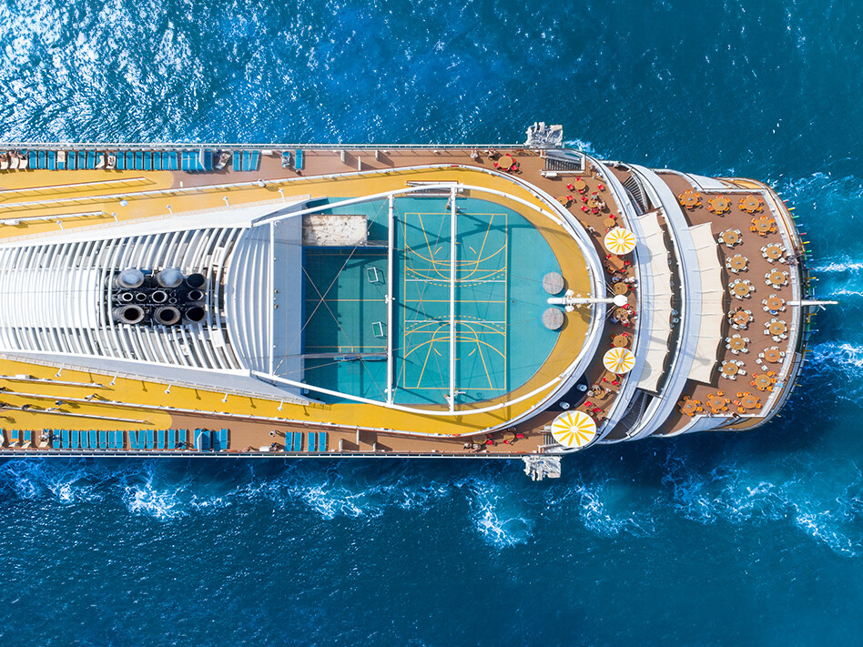 Aerial view of a cruise ship in blue water showing multiple decks with tables and chairs and a green playing court.