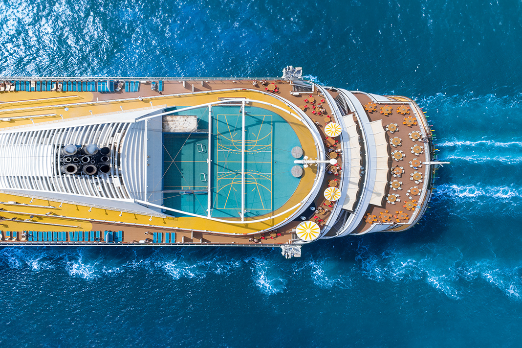 Aerial view of a cruise ship in blue water showing multiple decks with tables and chairs and a green playing court.