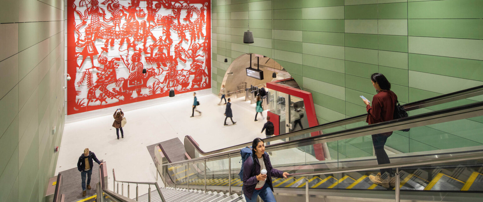 Concrete block walls with raised red sculptural mural on left. Right, arched hall with gates and ticket booth by escalators