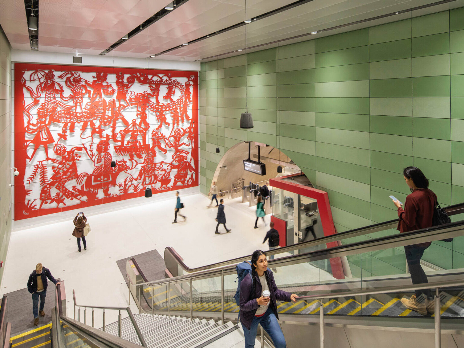 Concrete block walls with raised red sculptural mural on left. Right, arched hall with gates and ticket booth by escalators