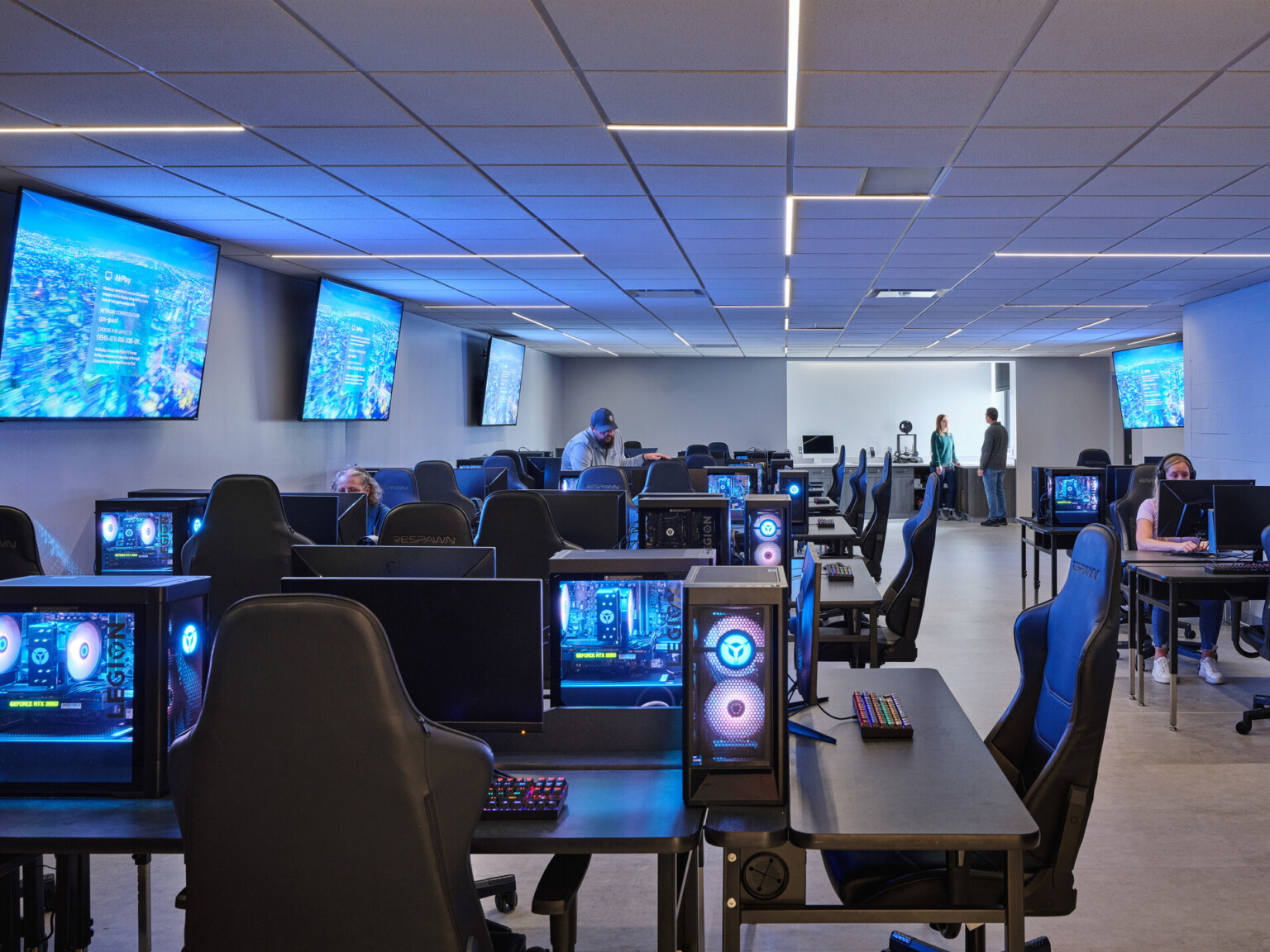 A modern computer lab classroom filled with computers lit from the inside at long tables and chairs, wall-mounted screens, and modern overhead lighting.