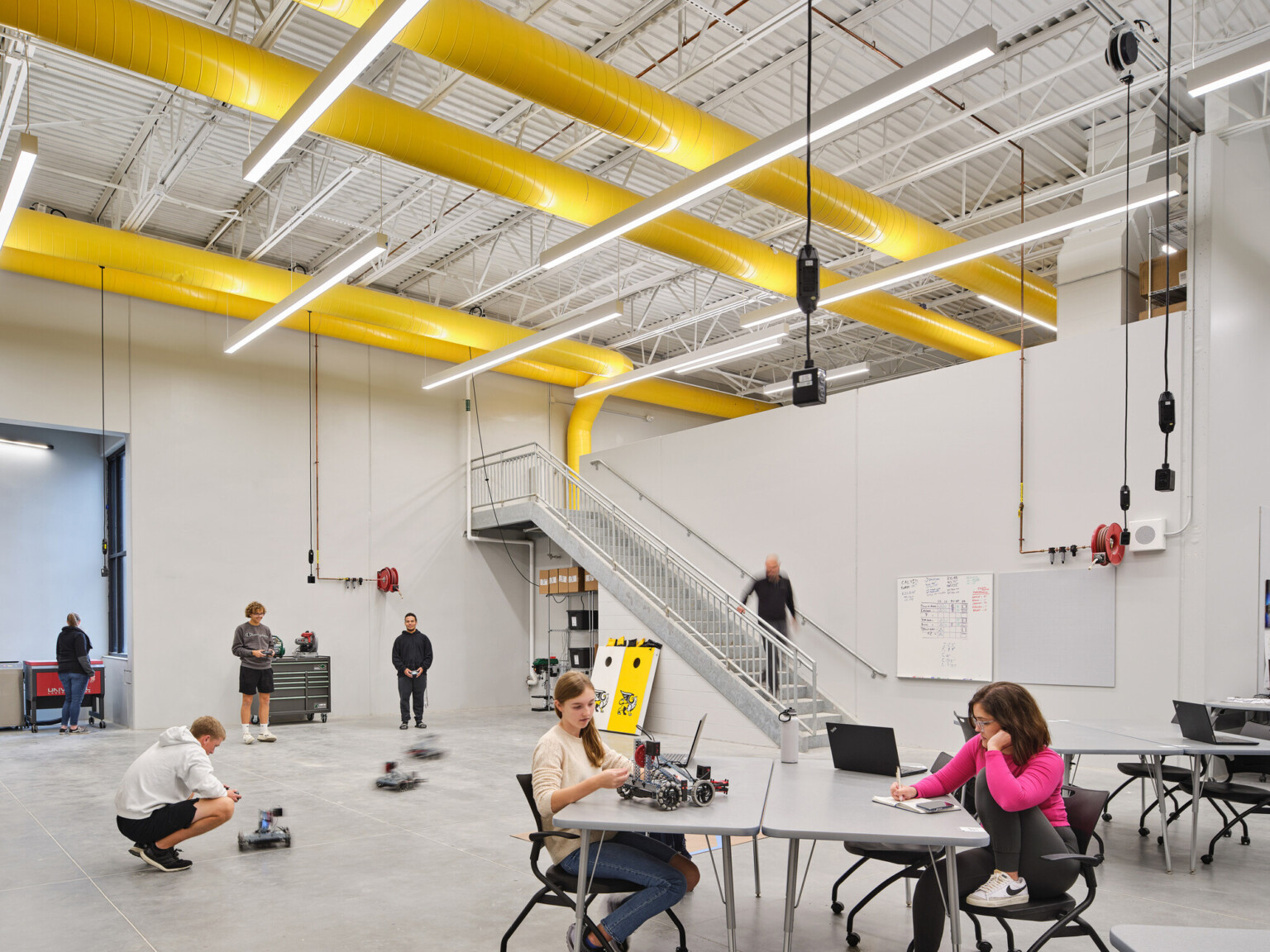 A spacious, industrial-style robotics lab with people working on projects at grey tables. Yellow ductwork in the ceiling.