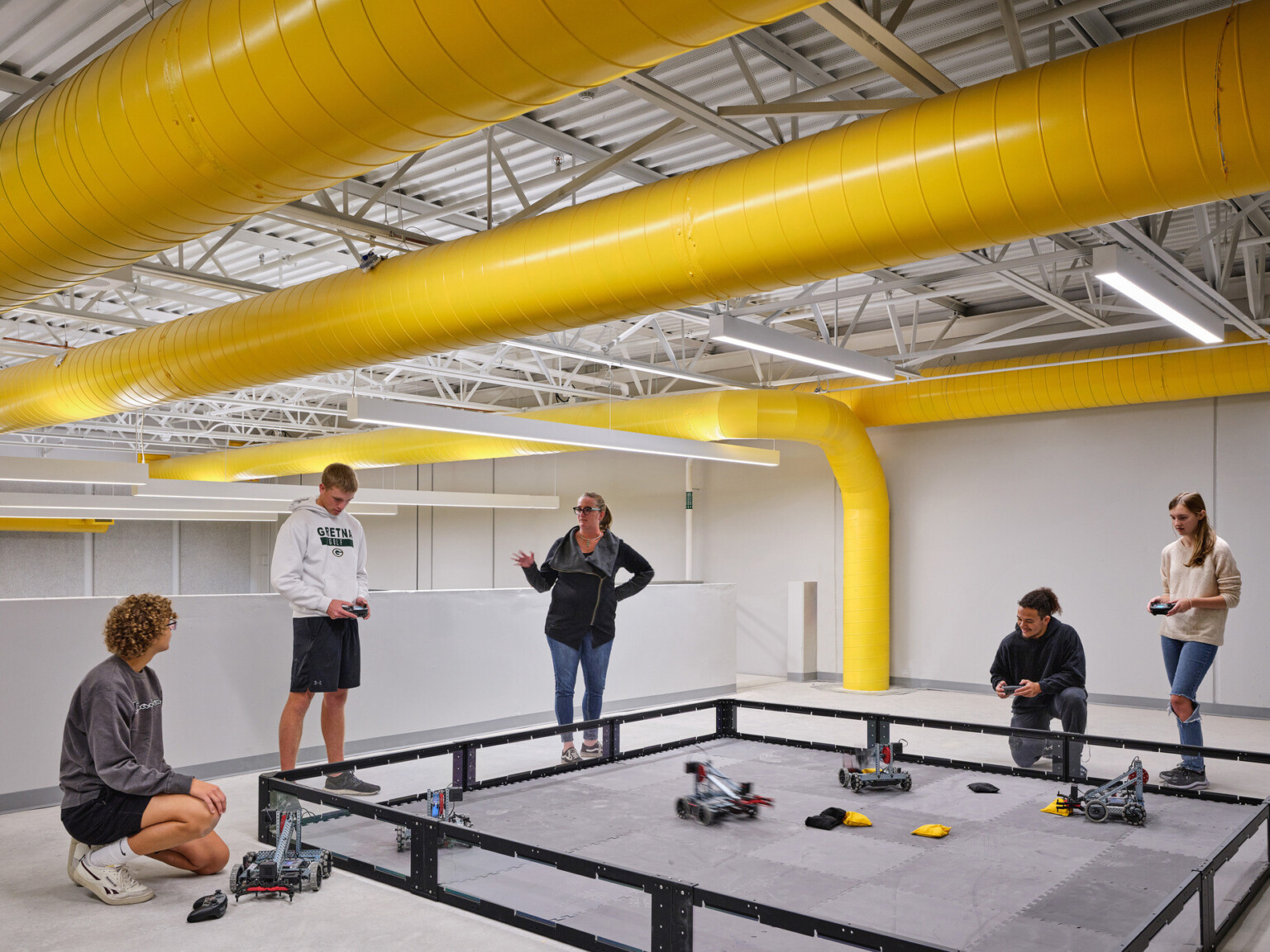 People in a room with yellow ceiling ducts, controlling small robots in a square arena on the floor.