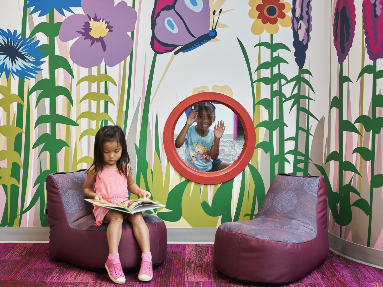 Children's reading area with colorful wall art of flowers and a butterfly, featuring a girl reading in a chair and another child peering through a window.