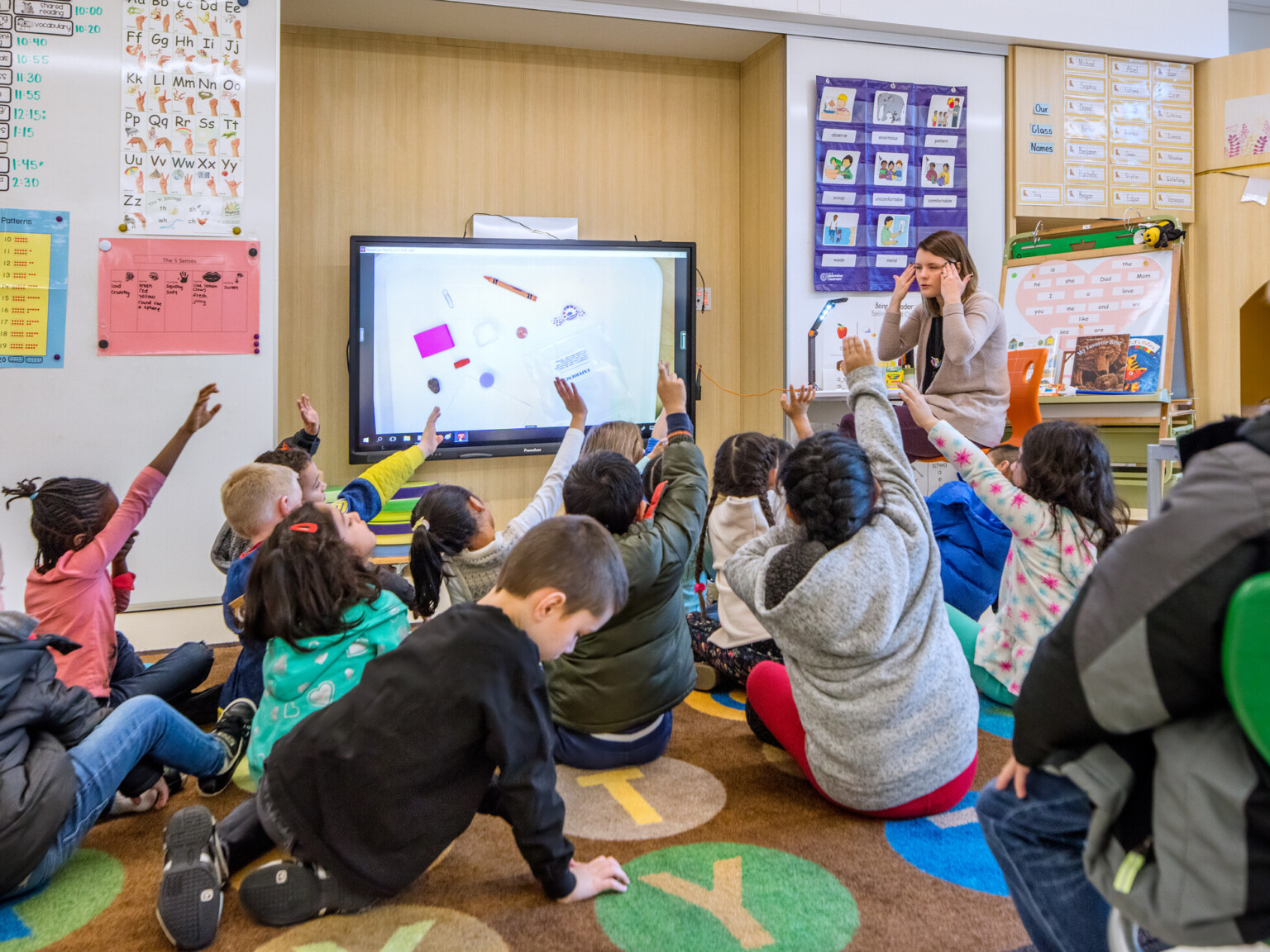 Children in a classroom sitting on the floor raising hands as a teacher sits in front of a smart board.