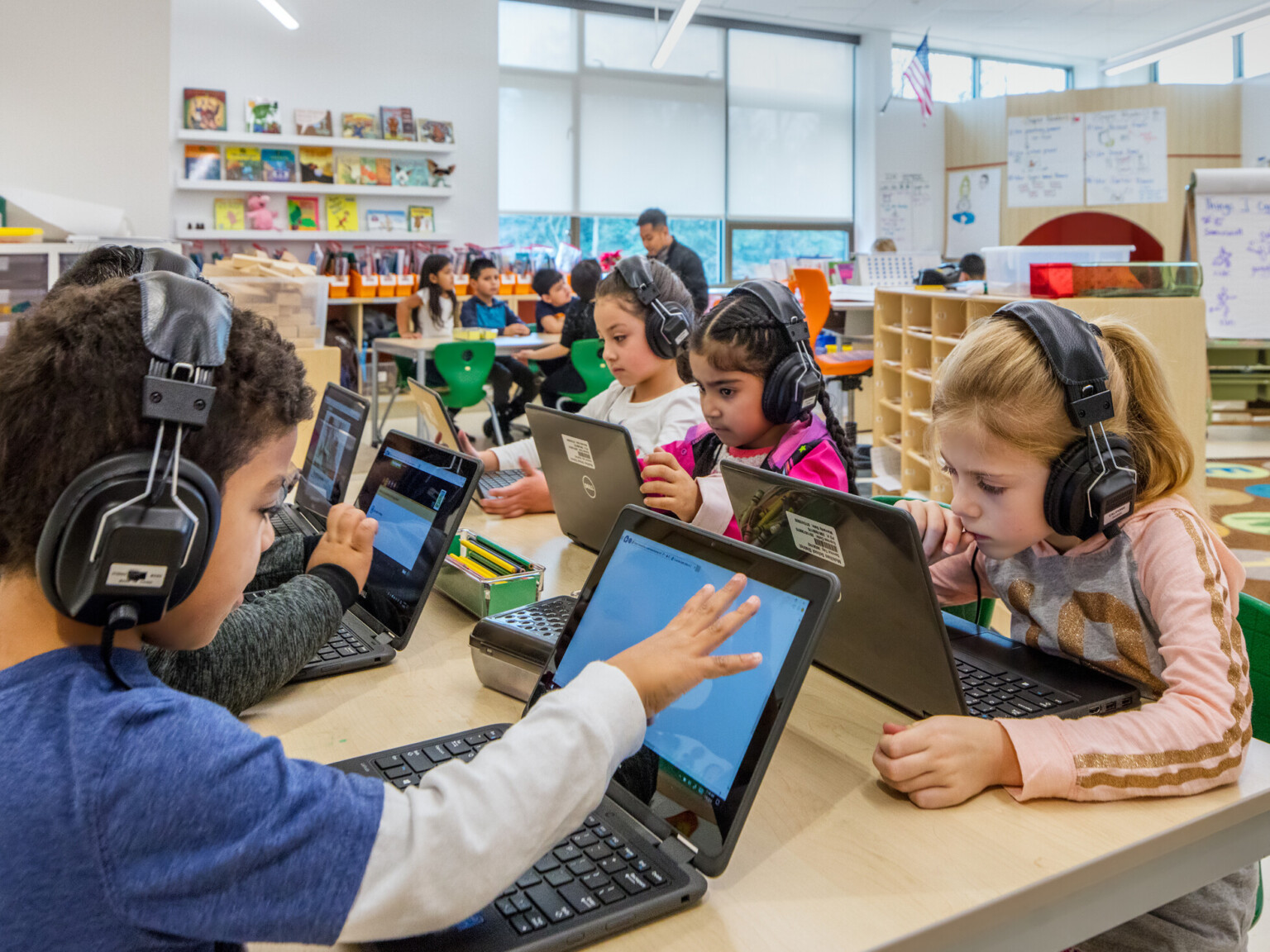 Group of children at a table using laptops in a classroom setting, wearing headphones.