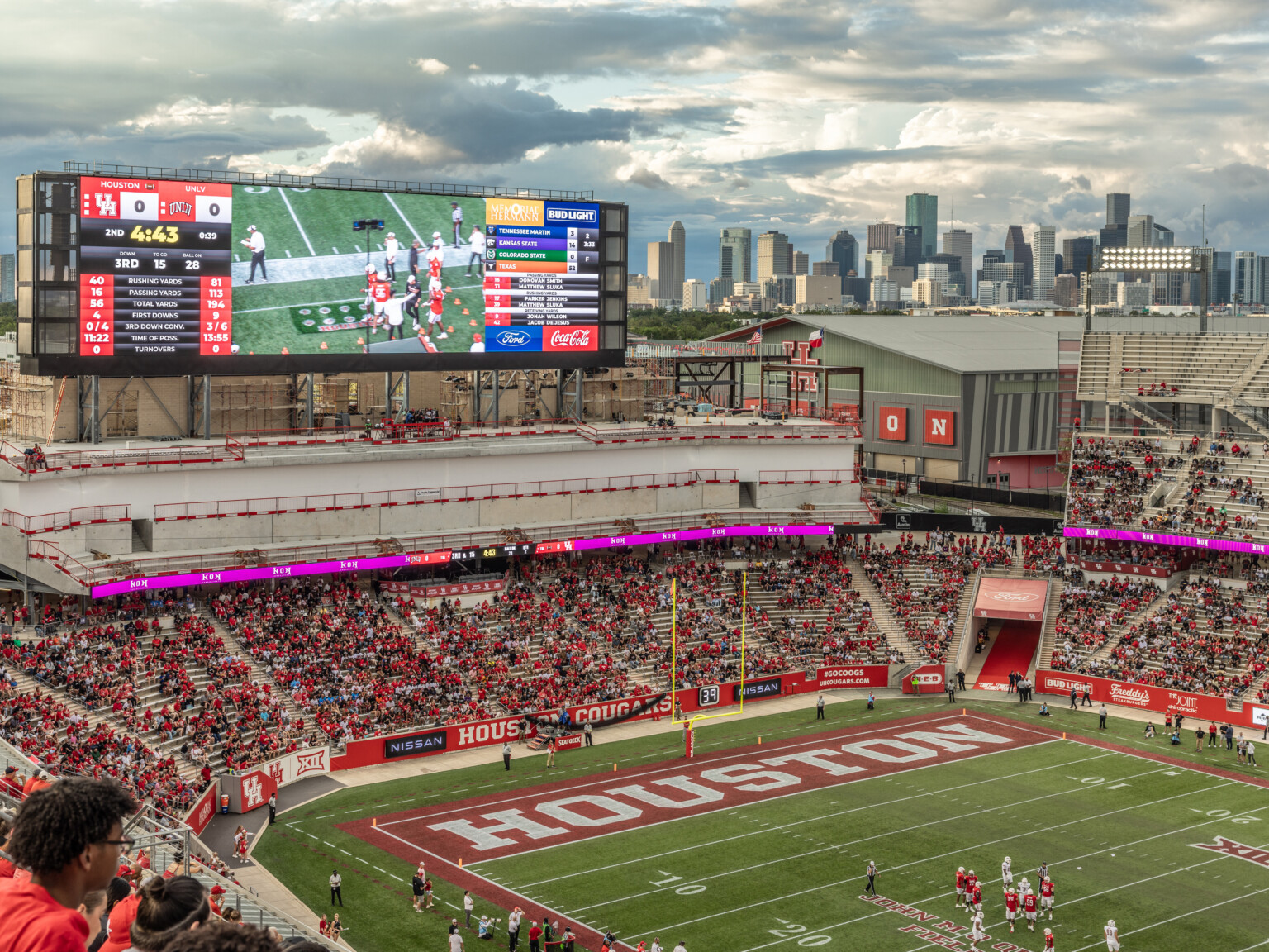 Football stadium filled with fans showing a large scoreboard with a view of skyscrapers in the background.