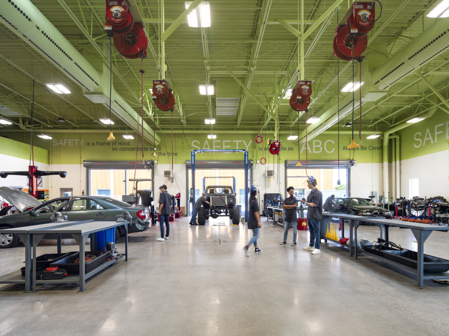 Automotive workshop classroom filled with cars, students engaged in tasks, tools on workbenches and red plugs hanging from the ceiling.