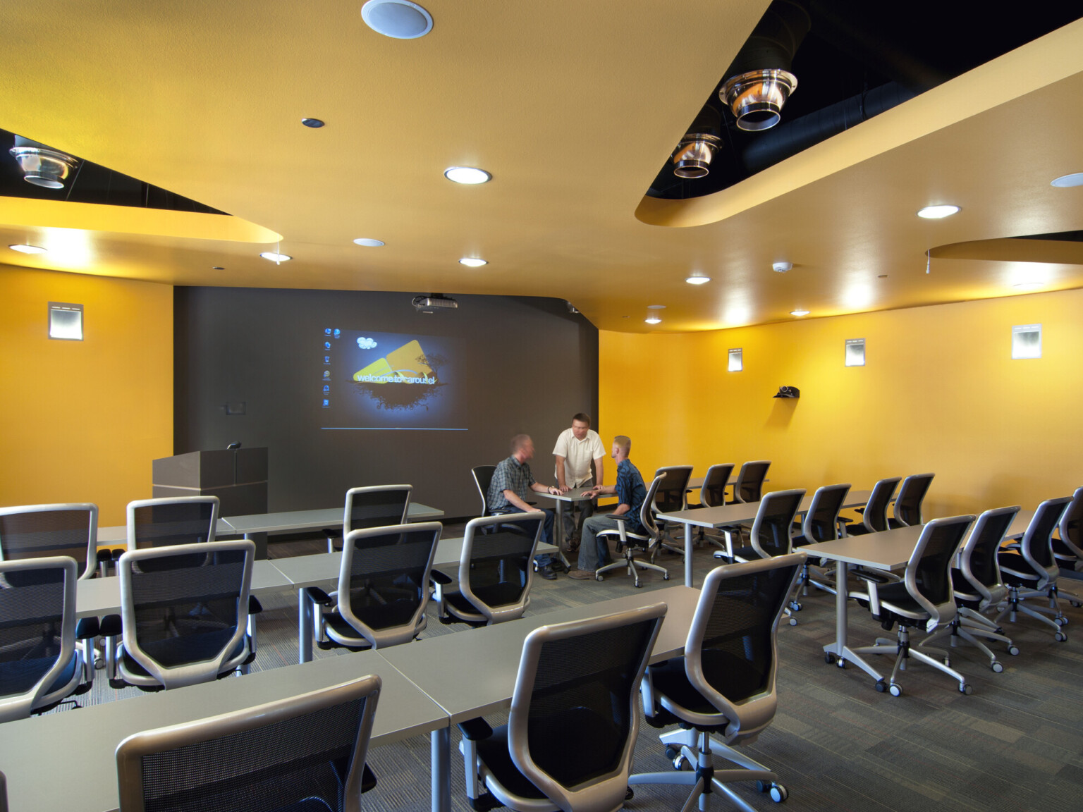 Modern classroom with yellow walls, grey chairs, and projector screen.