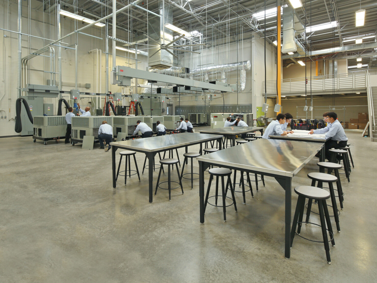 Industrial workshop classroom with metal tables and stools, people working on large machinery.